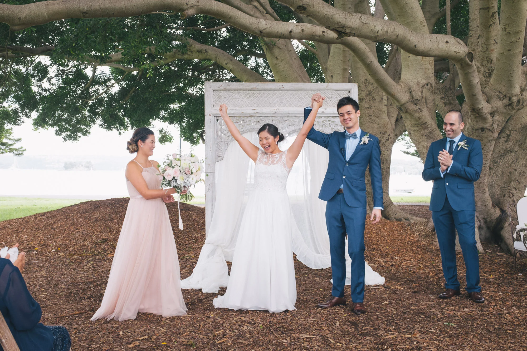 A wedding ceremony taking place outdoors under a large tree with a white decorative backdrop. The bride and groom are smiling, holding hands, and celebrating. The bride is wearing a white wedding dress, and the groom is dressed in a blue suit with a 