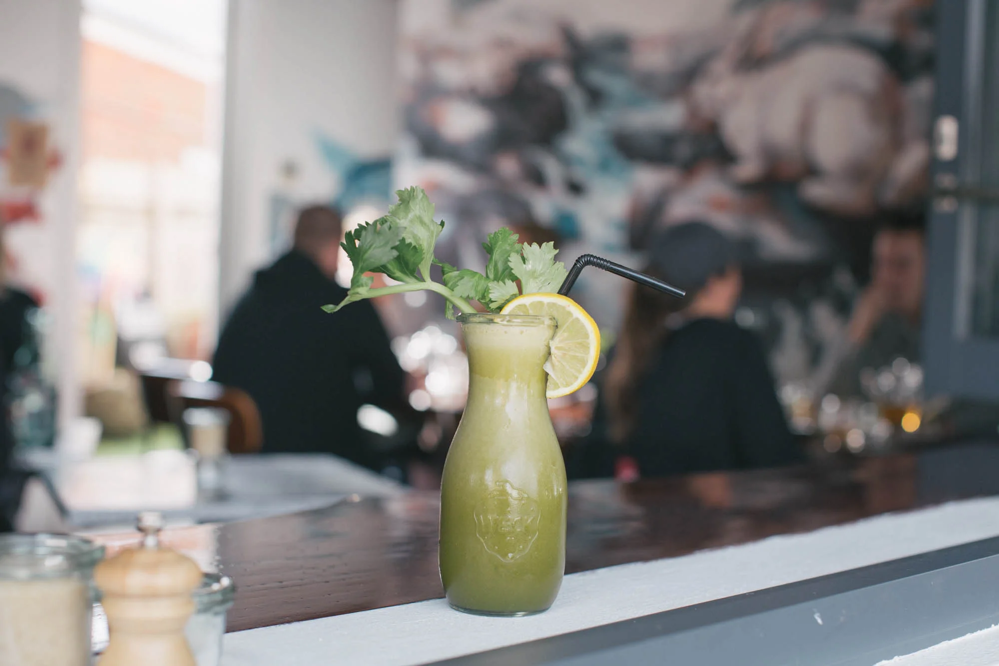 Green smoothie in a glass bottle with lemon slice and leafy garnish on a table in a café setting.