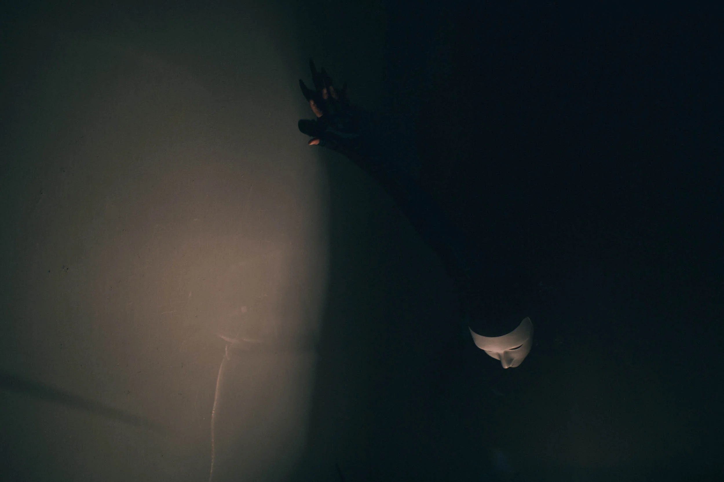 Dark scene with a figure in a white mask reaching towards a lit area.