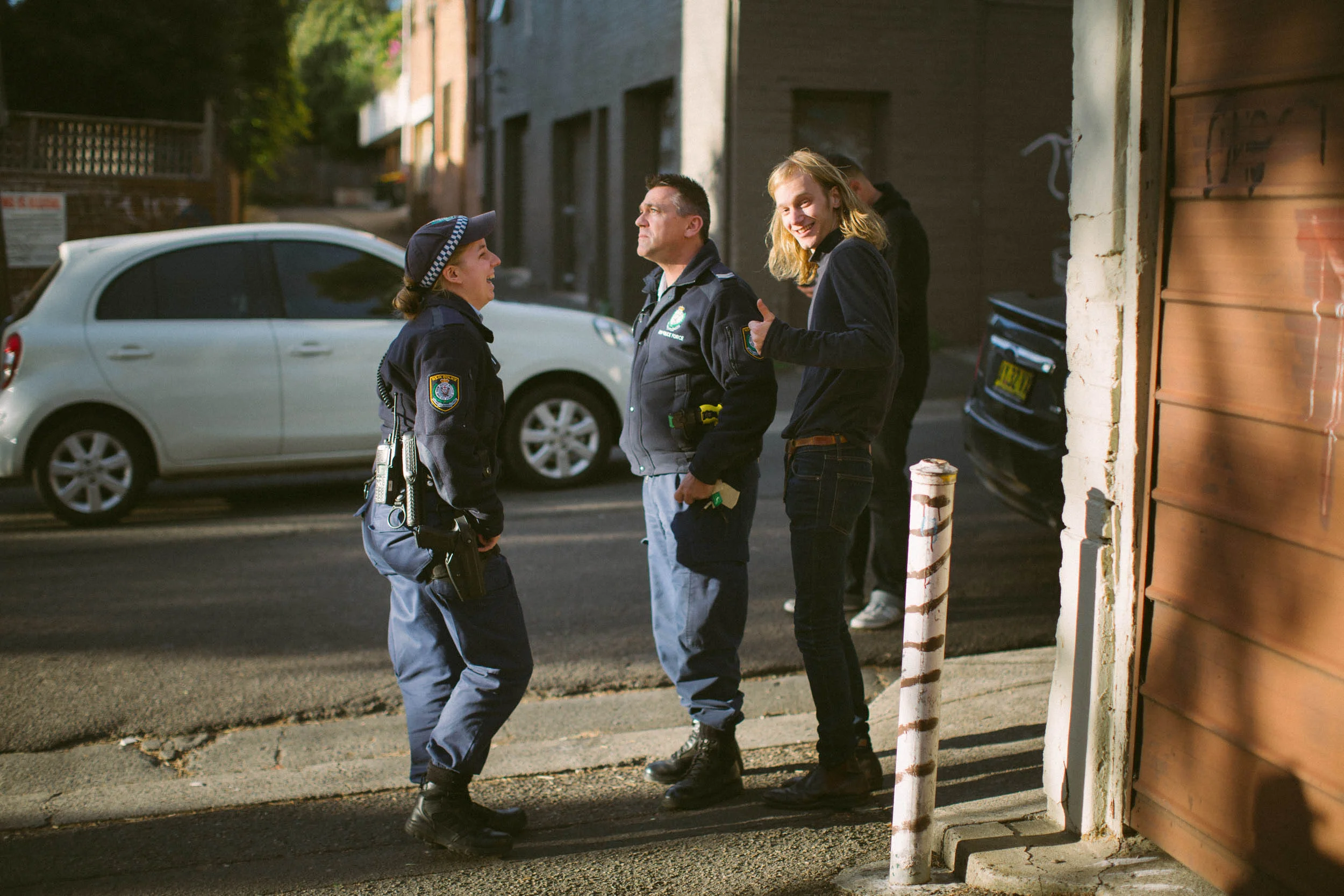 Two uniformed police officers and a smiling person standing on a street next to a parked white car.
