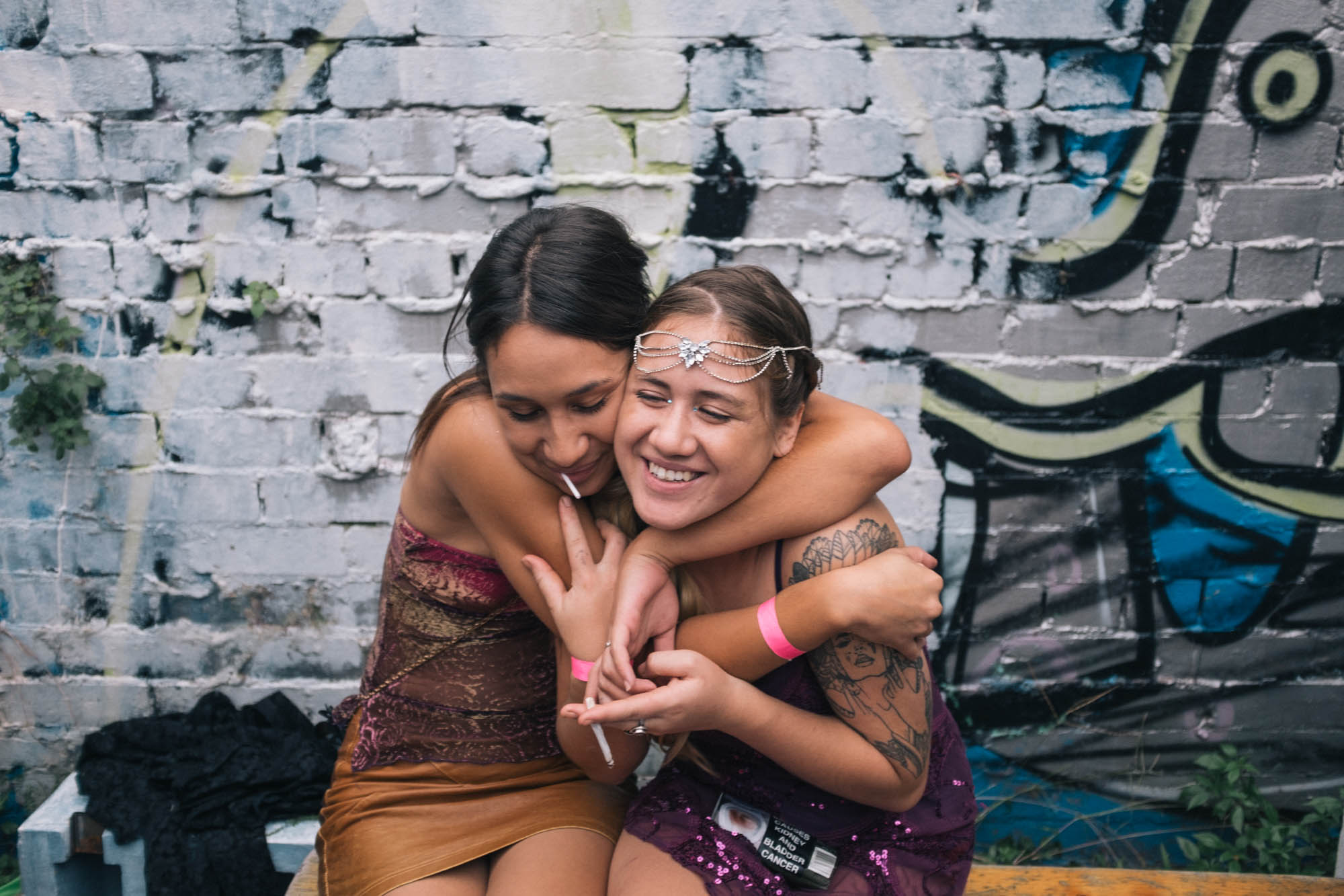 Two women hugging and smiling in front of a graffiti-covered brick wall.