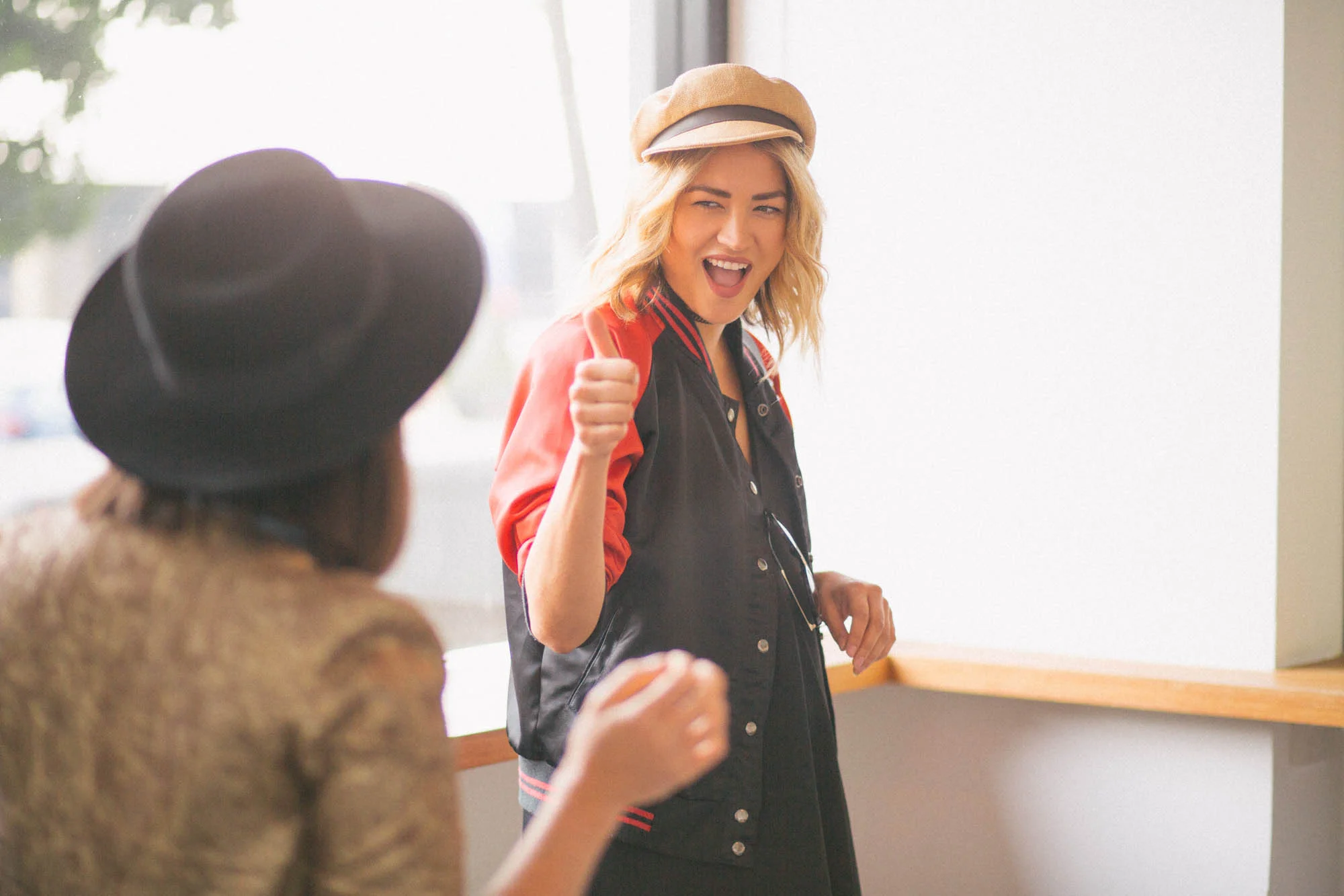 Two people interacting playfully indoors, one wearing a beige hat and red jacket, the other wearing a black hat.