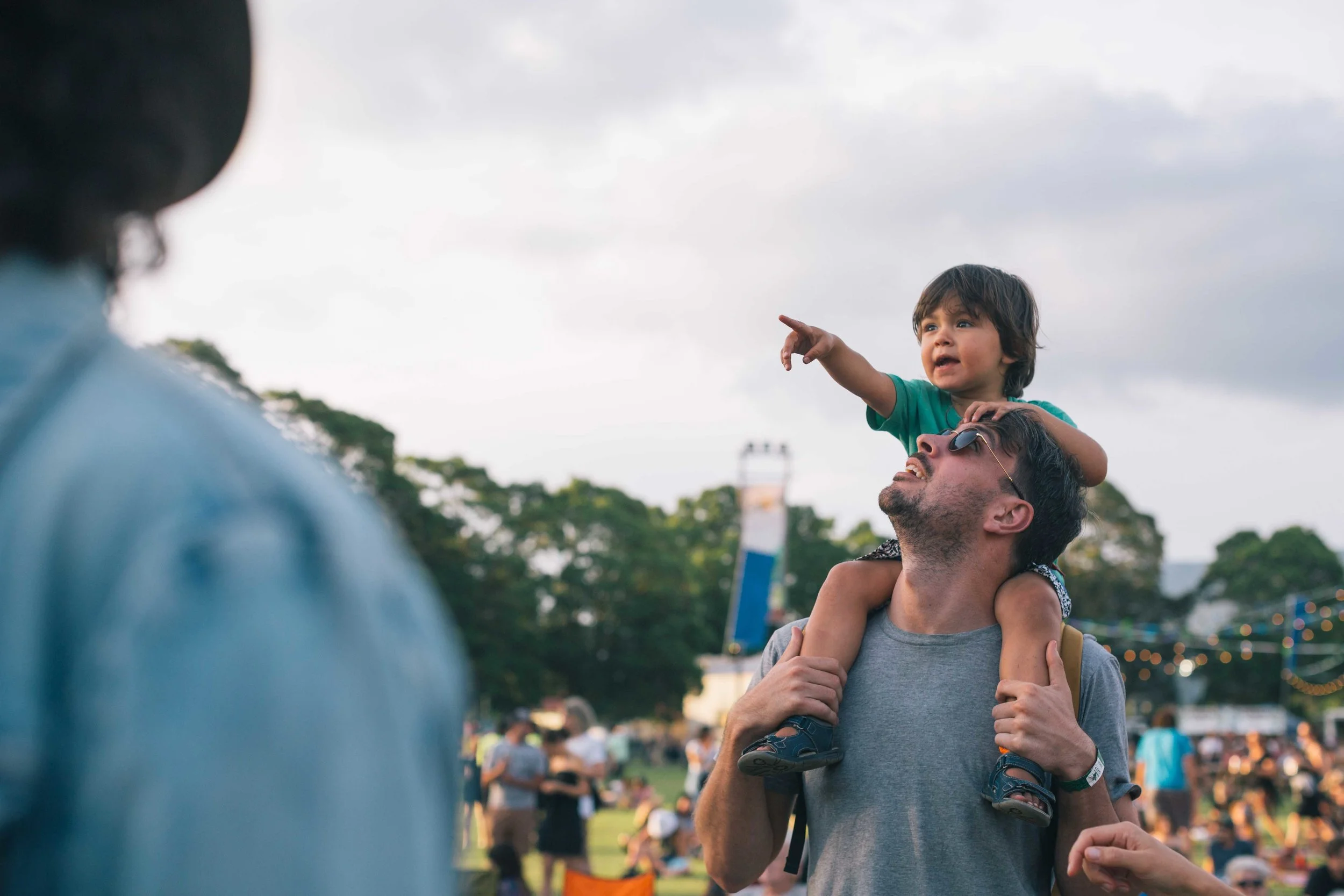 Child sitting on a man's shoulders at an outdoor event, pointing at something in the distance, with a crowd and trees in the background.
