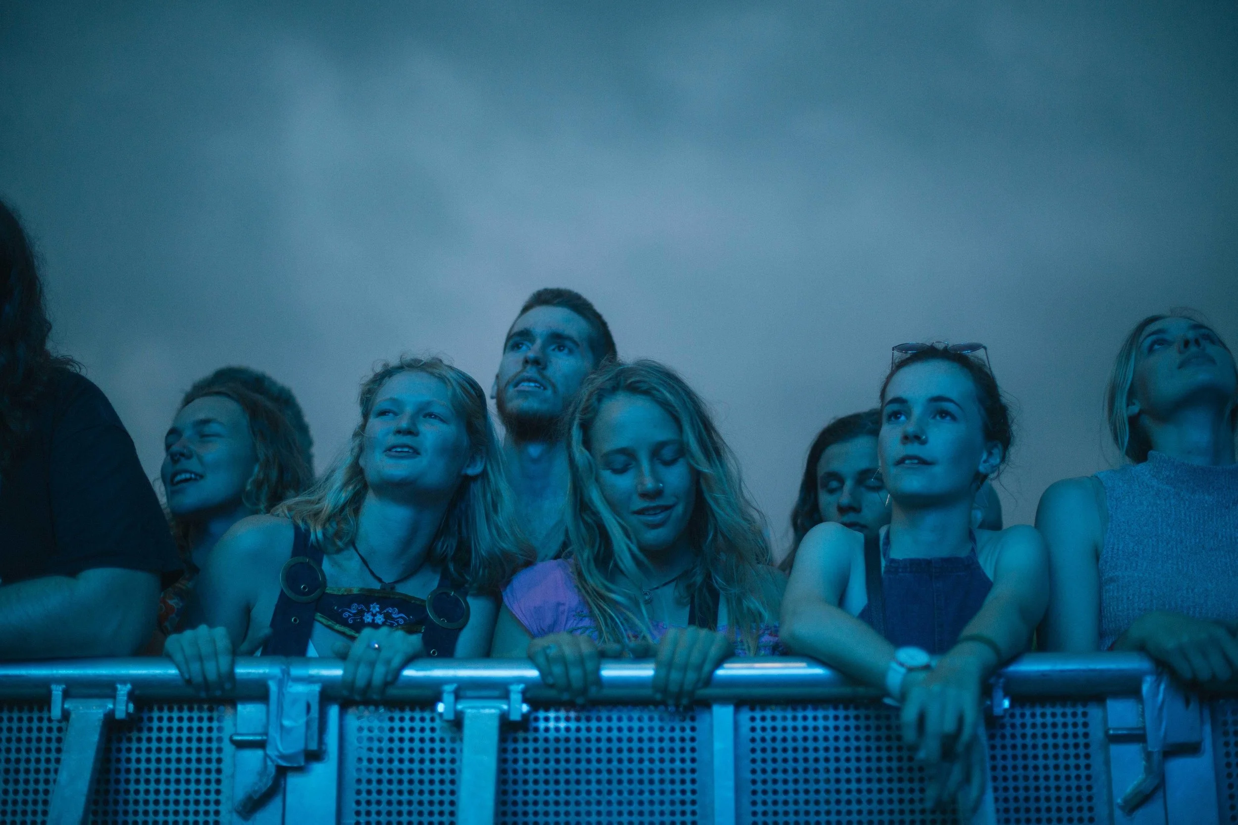 Group of young people at a music festival, standing at a barrier, illuminated by blue light, looking up towards the stage.