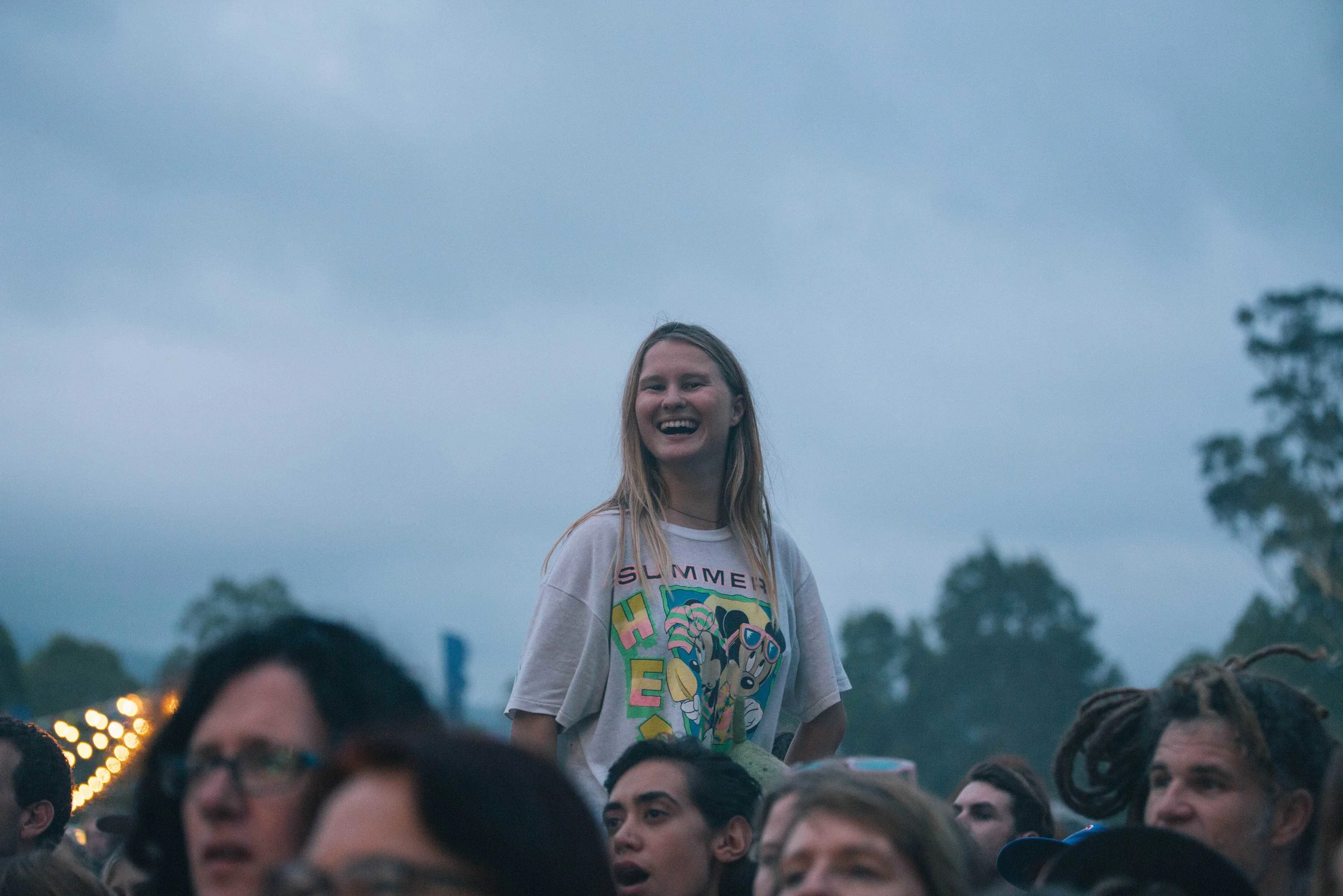 Crowd at outdoor concert, woman smiling standing on shoulders, evening sky in background.