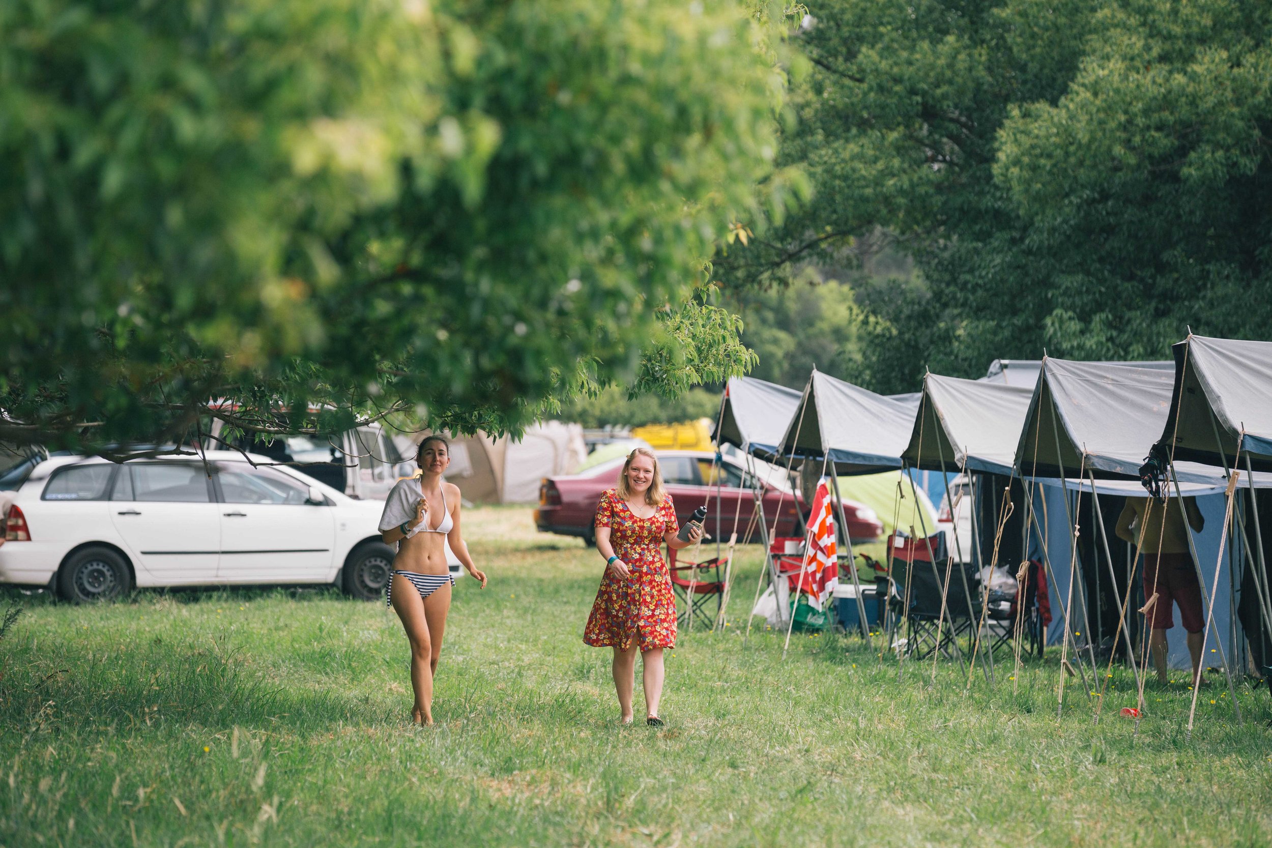 Two women at a campsite with tents and a car, surrounded by greenery.