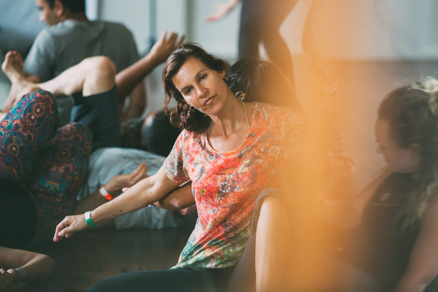 Group of people participating in a movement or dance workshop, with women seated in front and others in the background, some lying on the floor; warm, soft lighting.