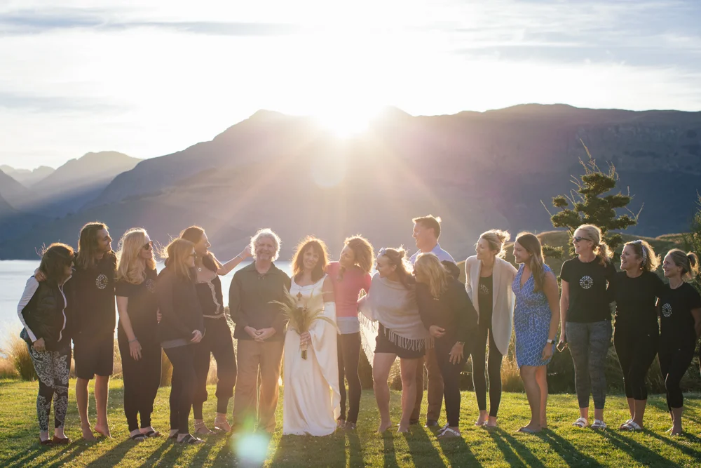 Group of people celebrating outdoors near a lake with mountains in the background during sunset.