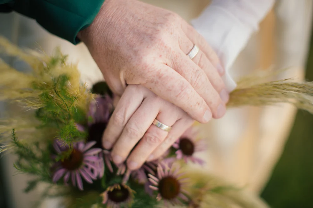 Close-up of two hands with wedding rings resting on a bouquet of purple flowers and dried grasses.