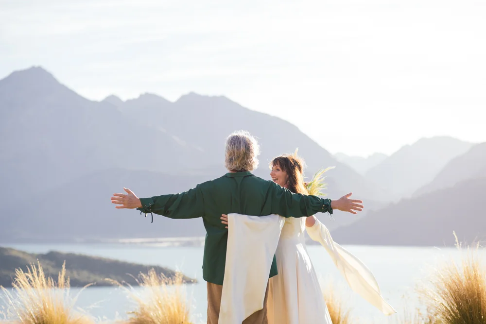 A man and woman in wedding attire embracing with arms outstretched outdoors near a mountain lake during sunset.
