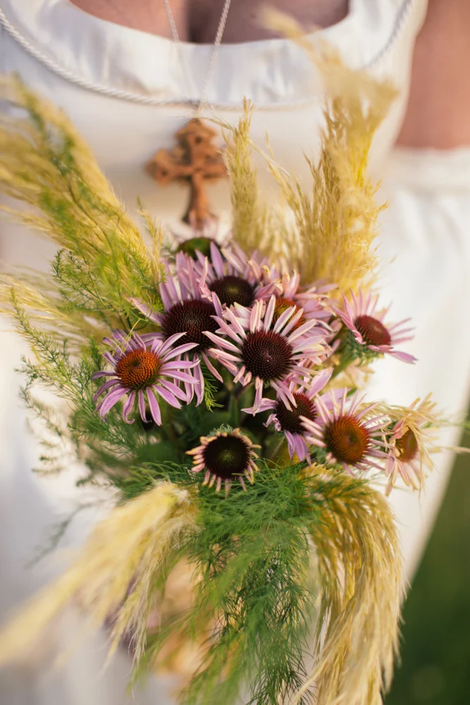 A person in a white dress holding a bouquet of purple coneflowers and green foliage with a wooden cross necklace around their neck.
