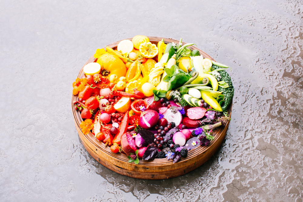 Colorful fruit and vegetable platter on a round wooden tray, featuring strawberries, oranges, peppers, greens, and purple root vegetables.