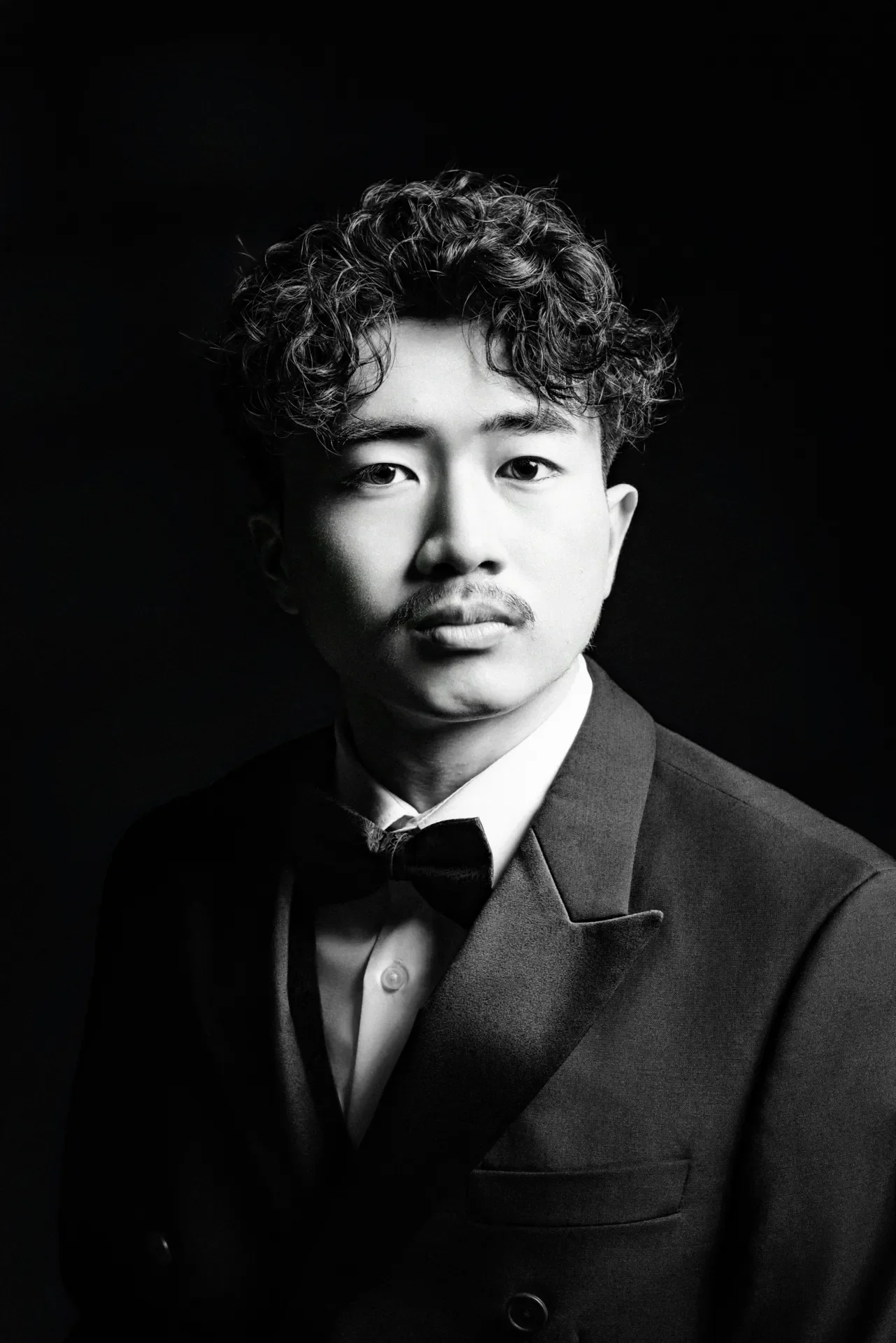 Black and white portrait of a young man wearing a tuxedo with a bow tie, with curly hair and a mustache, against a dark background.
