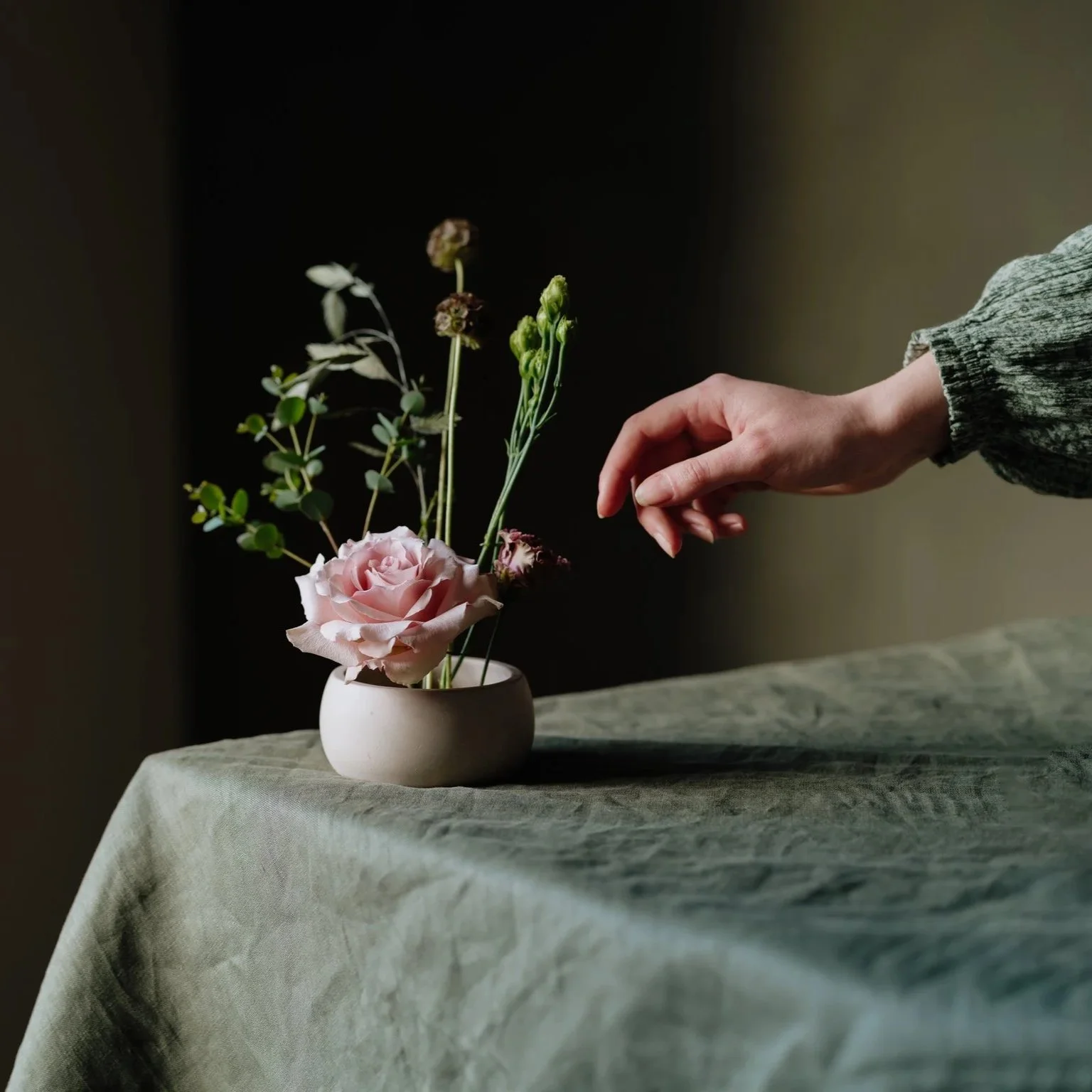 A female hand reaching towards an ikebana flower arrangement in a bowl on top of a table covered with a green tablecloth