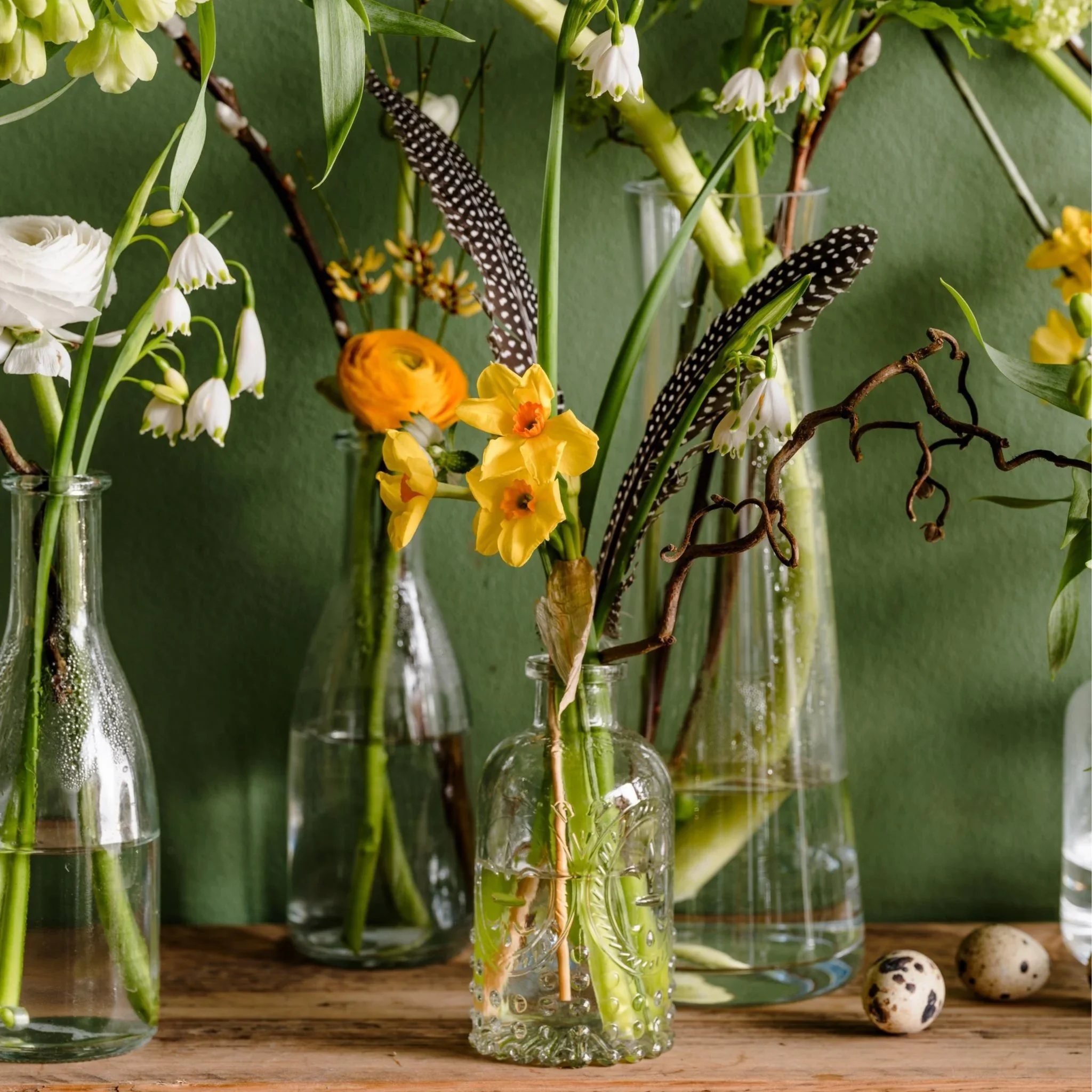 Yellow and white flowers in glass vases against a green backdrop with feathers and small speckled eggs
