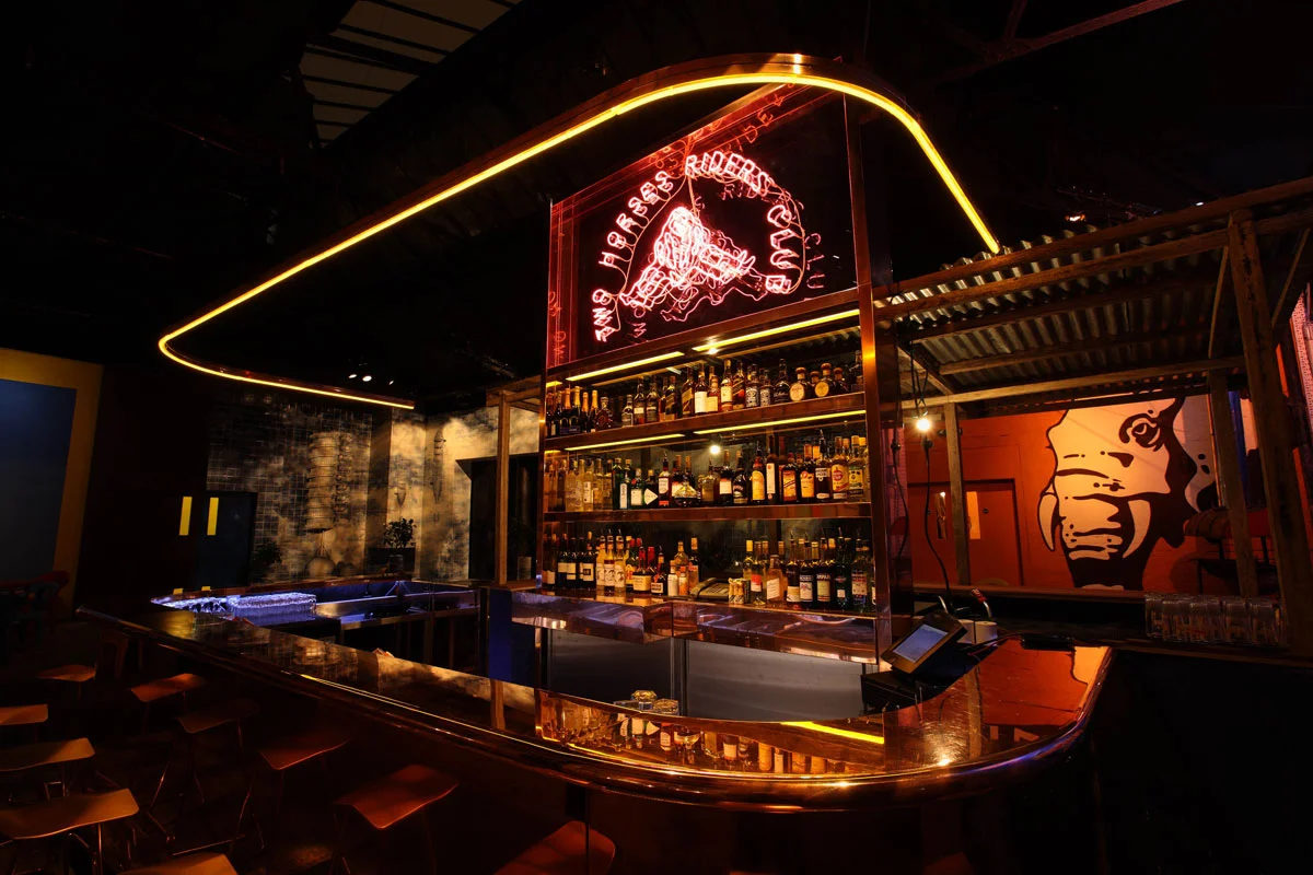 The Courtyard Bar was at the heart of the Double Club experience. The space was divided into four slices: two western and two Congolese, and was covered with a glass roof lending it a special outdoor feeling. The Congolese bar boasts imported wood and corrugated iron from Kinshasa. The Western bar, by contrast, is a solid copper monoblock lit up by neon signage announcing the Two Horses Riders Club — for people who enjoy riding two horses simultaneously.