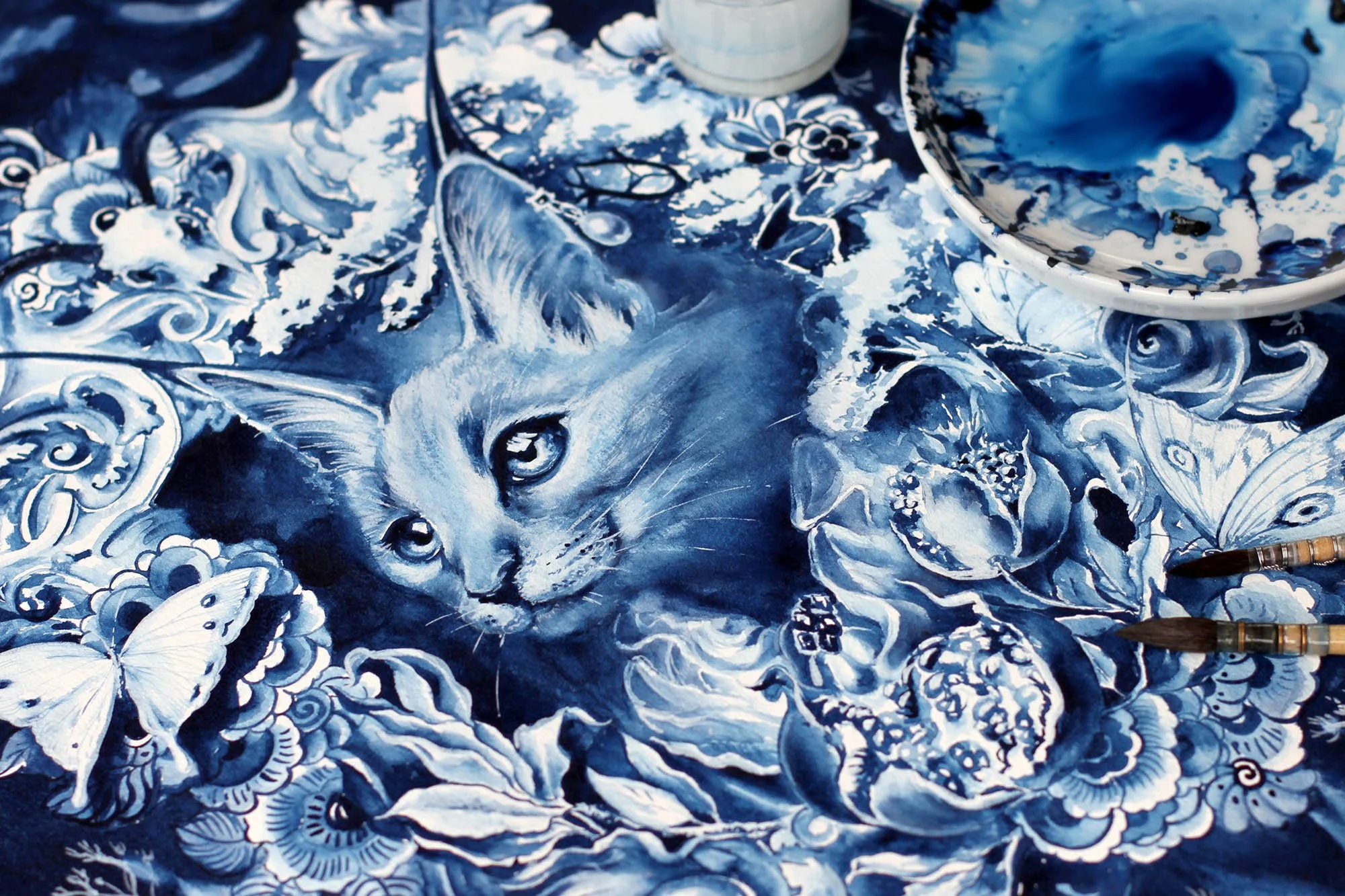 Close-up of a Persephone cat painting in progress, showing watercolor and gouache details, pomegranates, butterflies, and blue-and-white Delft-inspired ornamentation.