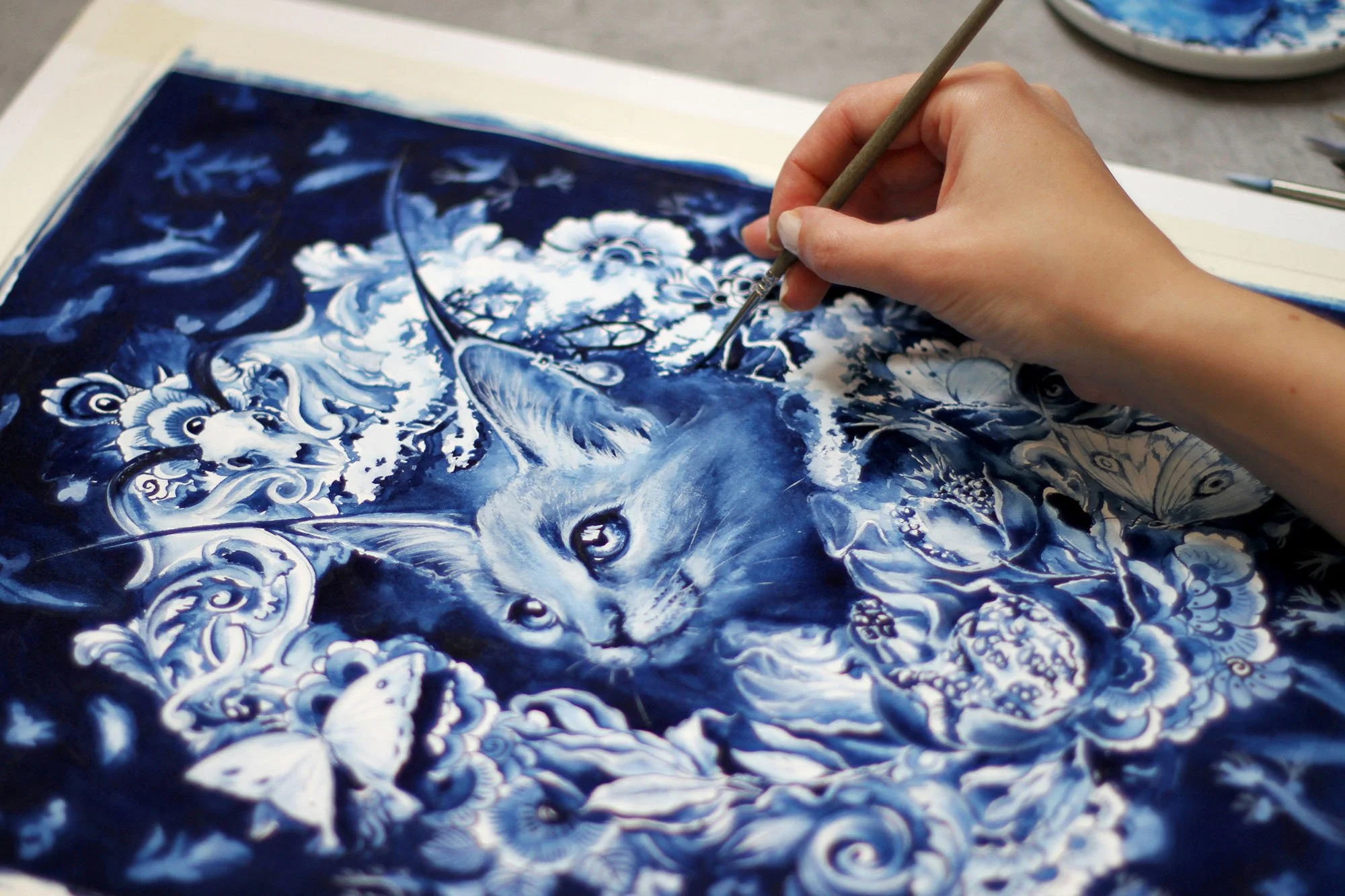 Artist painting the Persephone artwork with a fine brush, developing details of the mythological cat portrait surrounded by pomegranates, flowers, and Delft Blue decorative motifs.