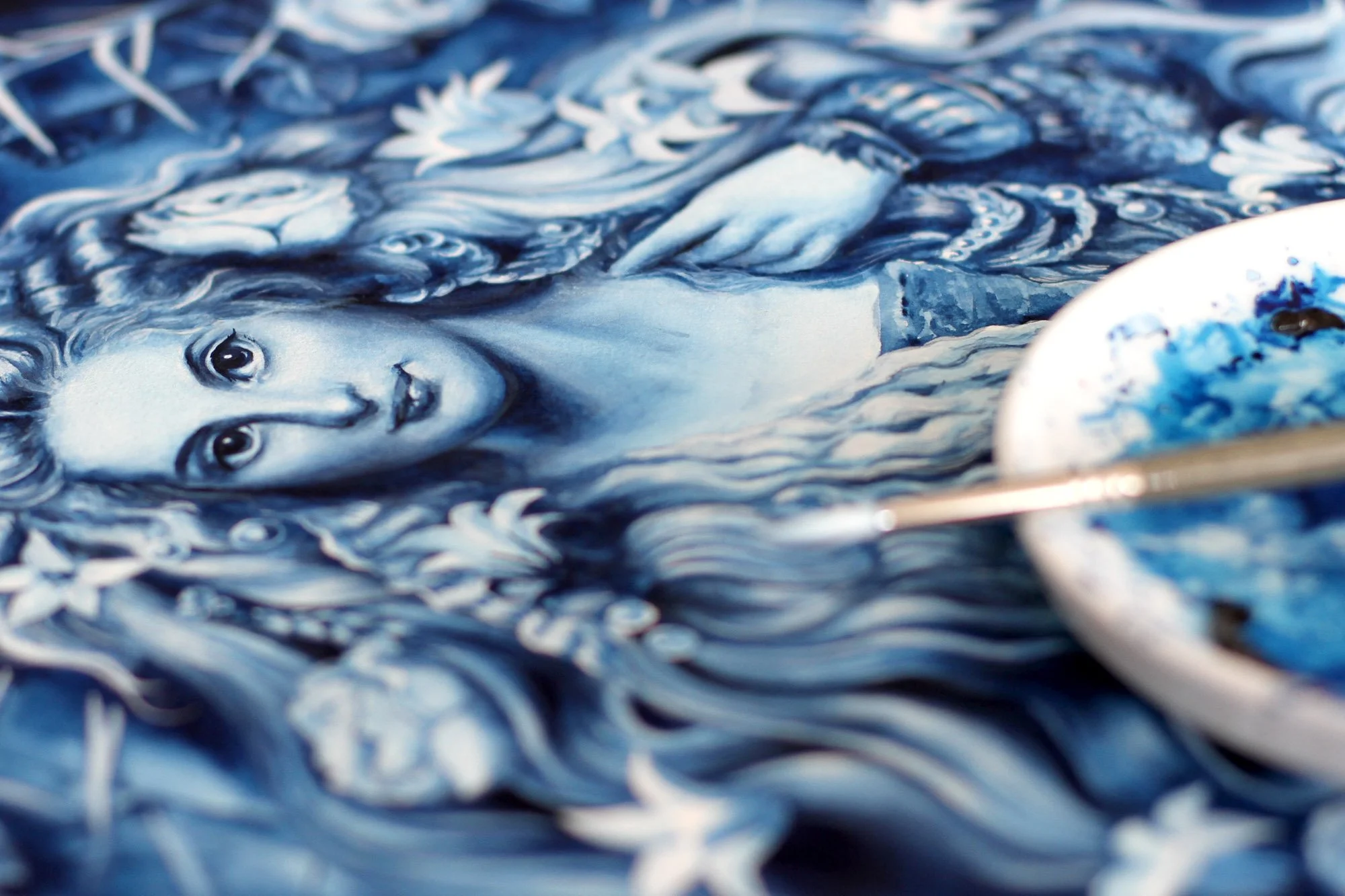 Process detail with brush and palette Close-up process photo of Romance of the Rose, showing the blue-and-white painted figure with floral details, thorny branches, a brush and blue pigment palette.