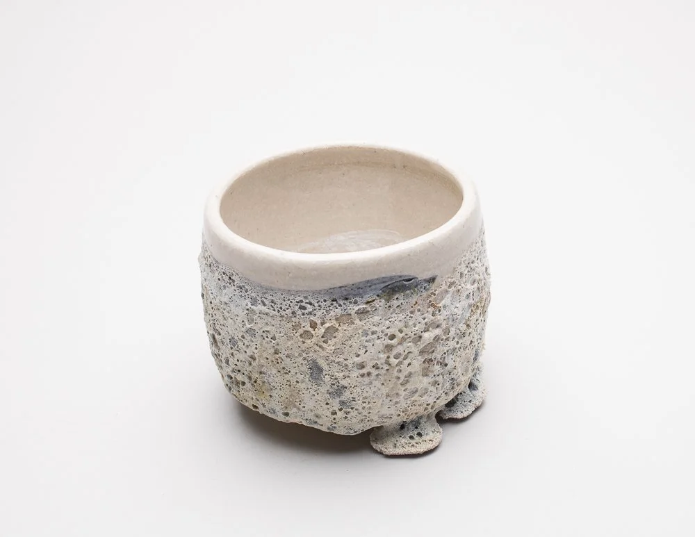 A handmade ceramic cup with a smooth white upper section and a textured, porous lower section resembling natural rock formations. The design blends modern minimalism with an organic, rustic aesthetic.