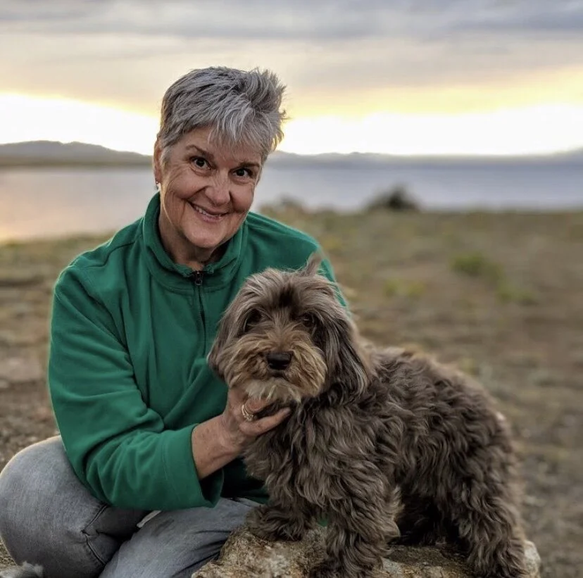 A woman with short gray hair and a green jacket sitting outdoors on a rocky surface, smiling at the camera, with a fluffy brown dog sitting on a rock in front of her near a body of water at sunset.