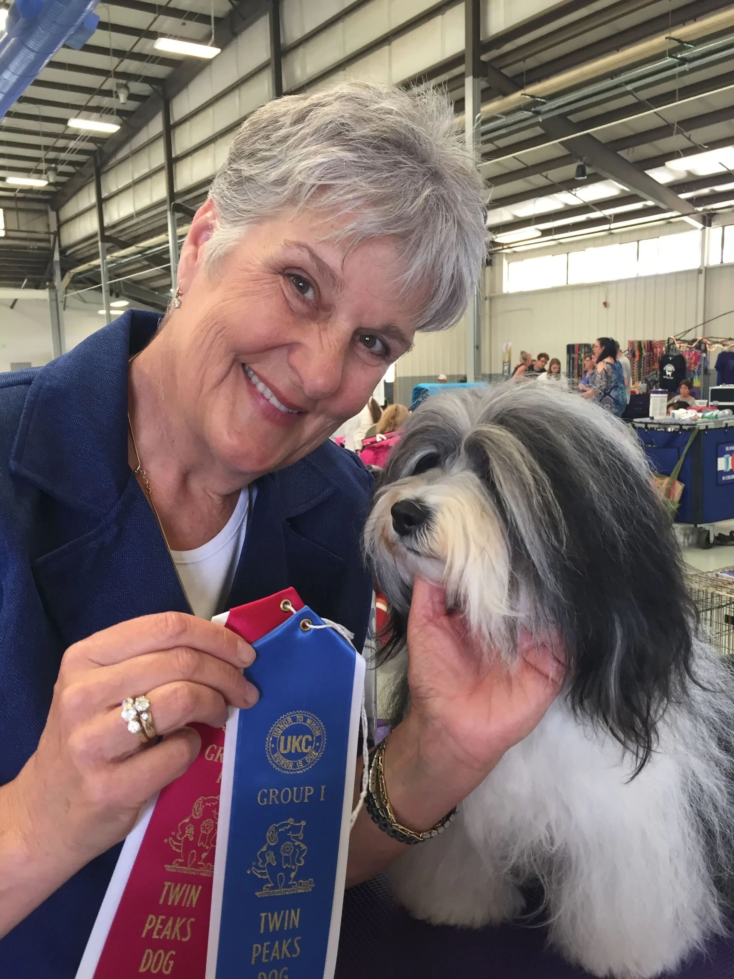 A woman with short gray hair holding a fluffy, long-haired dog at a dog show, with ribbons on display.