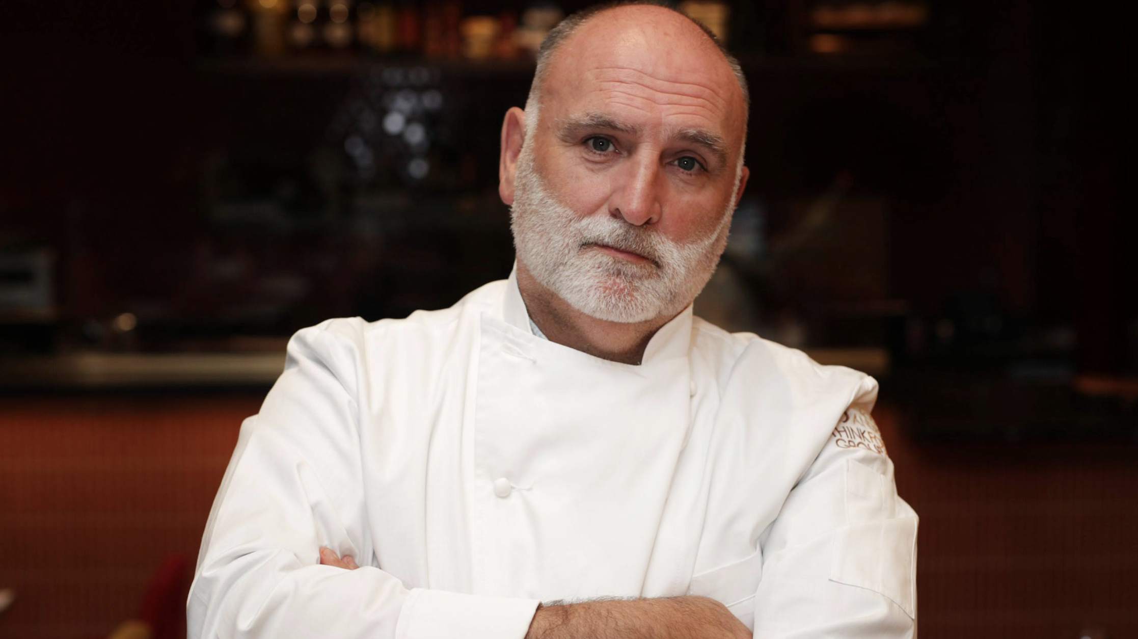 A professional chef with a white beard and bald head, wearing a white chef's coat, standing in a kitchen with a blurred background of shelves and kitchen equipment.