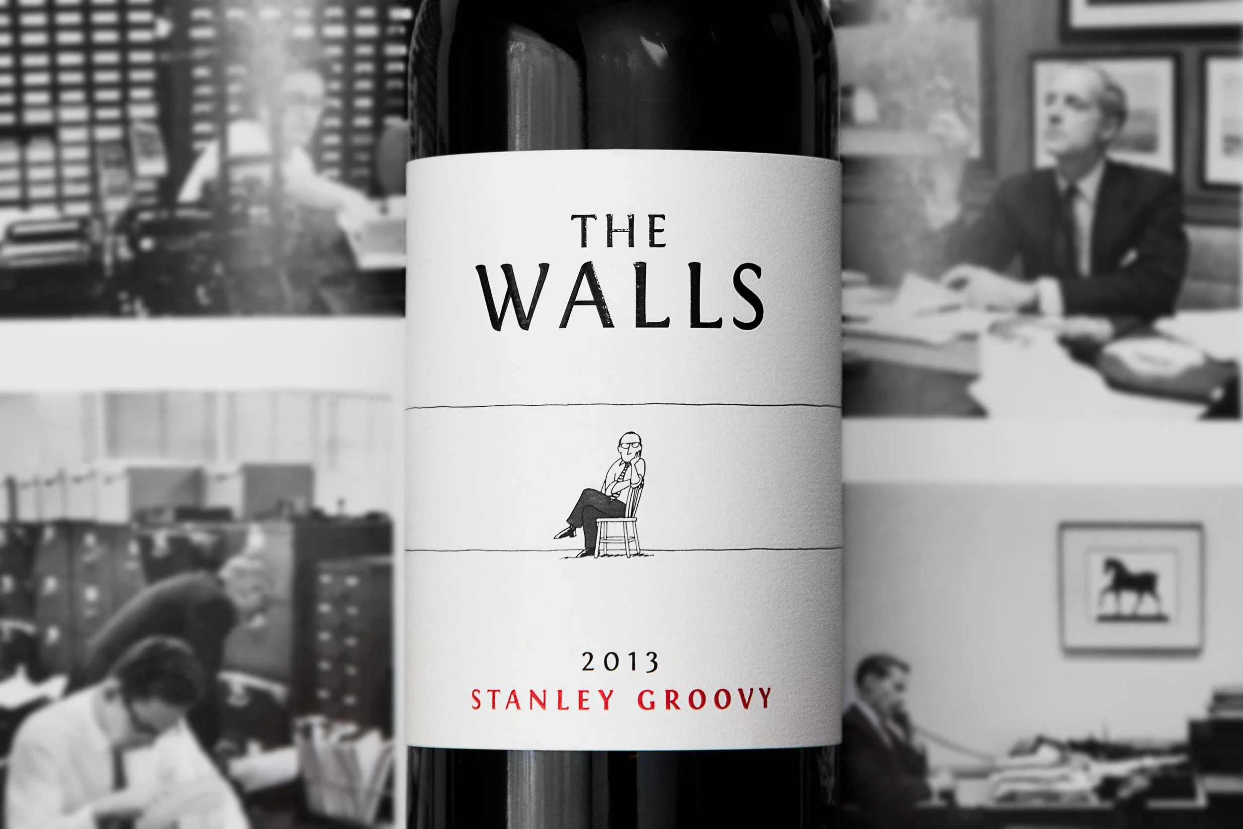 A bottle of red wine labeled 'The Walls,' featuring a minimalist illustration of a person sitting on a chair. The label indicates the wine is from 2013 and made by Stanley Groovy. The background shows black and white photographs of people in an office or meeting setting.