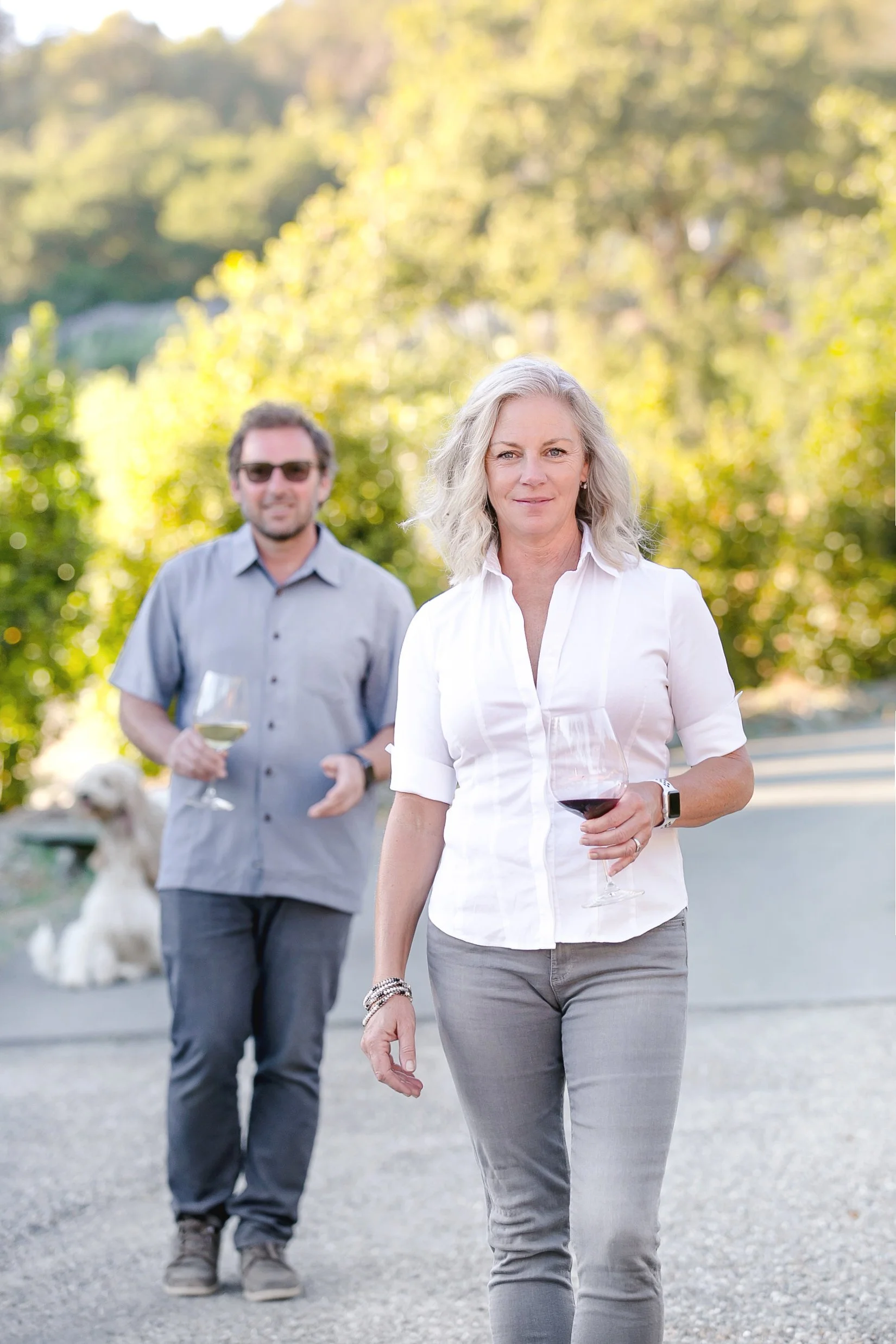A woman with blonde hair holding a glass of red wine, walking outdoors on a sunny day, with a man in the background holding a glass of white wine and a dog sitting nearby.