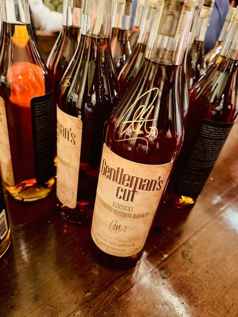 A group of bottles of Gentleman’s Cut Kentucky bourbon whiskey on a wooden surface, with prominent label and signature visible.