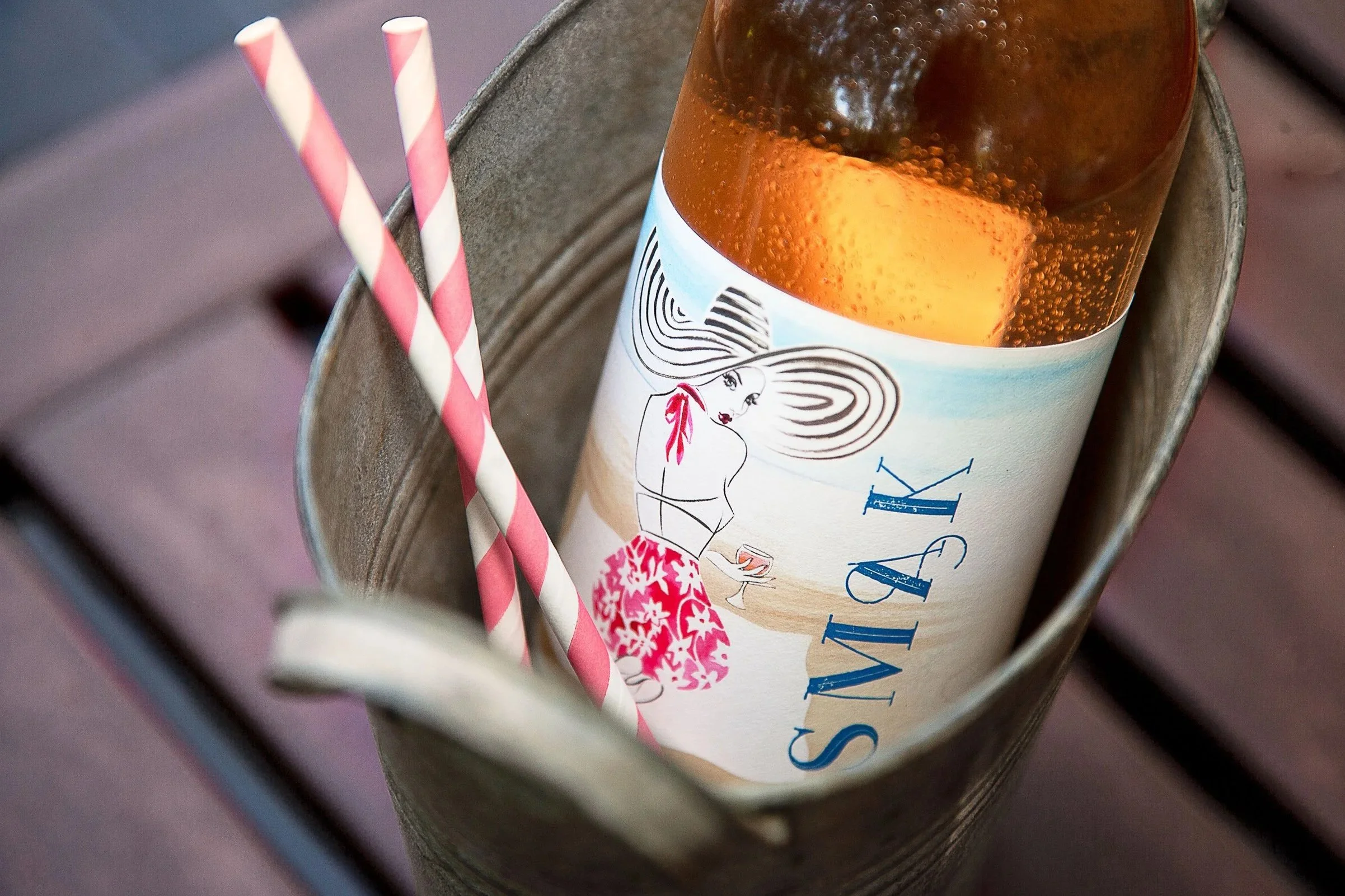 A close-up of a glass of soda with bubbles, placed inside a metal container. There are two striped pink and white paper straws leaning against the glass. The drink's label features an illustration of a woman with long hair and the word 'VIVA' in blue