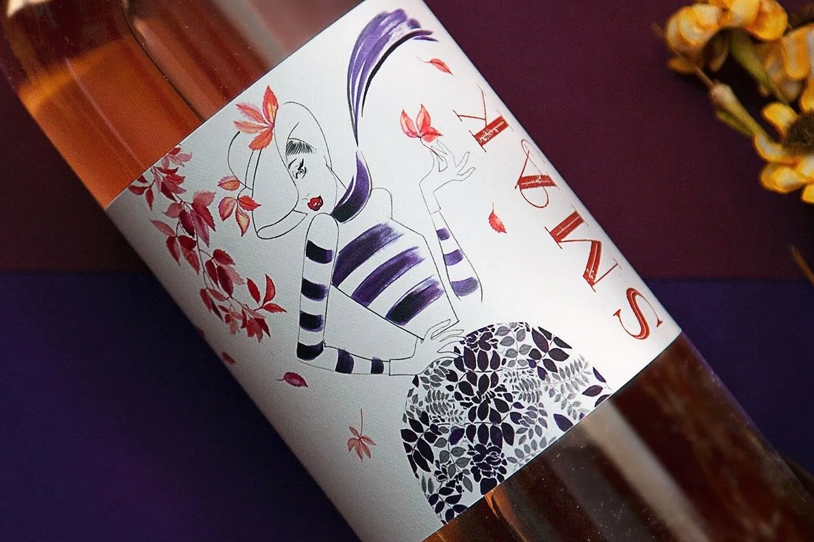 A wine bottle with a label featuring a stylized illustration of a woman with long hair wearing a striped shirt, surrounded by red leaves and a decorative black and white circular pattern. The label includes the text "SWA".