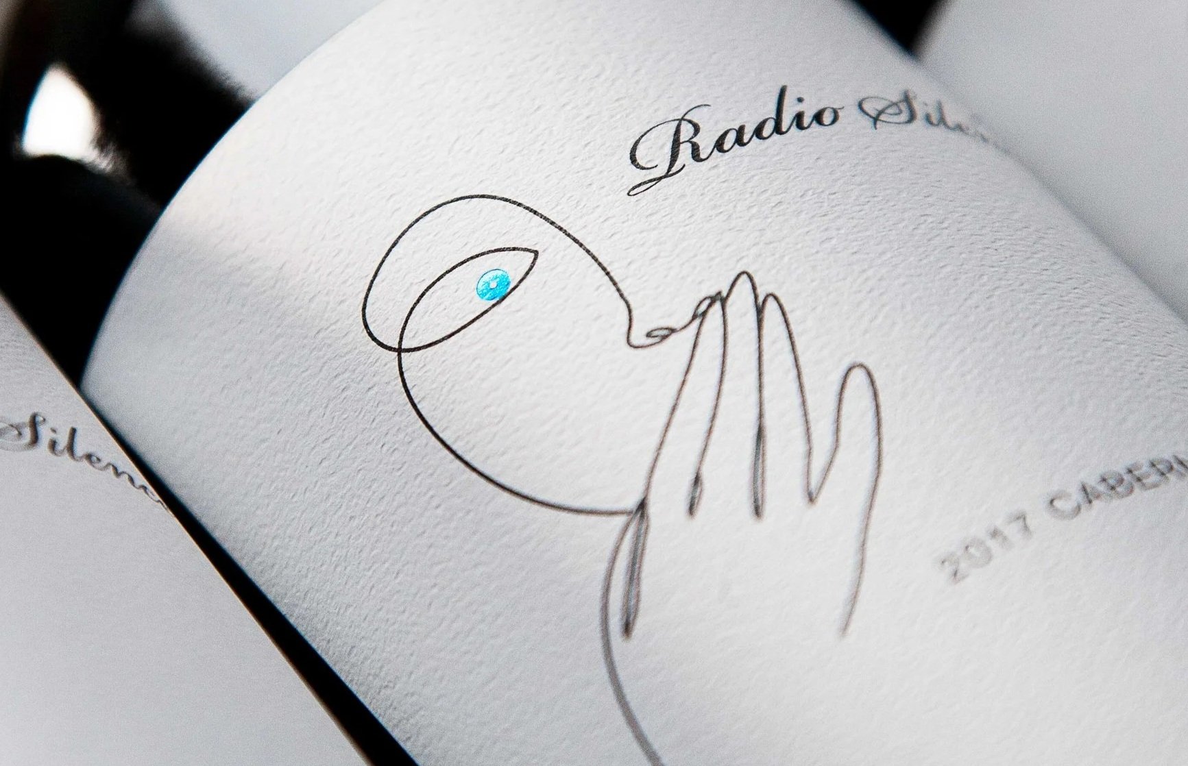 A piece of paper with a minimalist line drawing of a hand holding a glass. The paper has the words 'Radio Sil' written in elegant cursive, and there is a small blue dot on the glass.