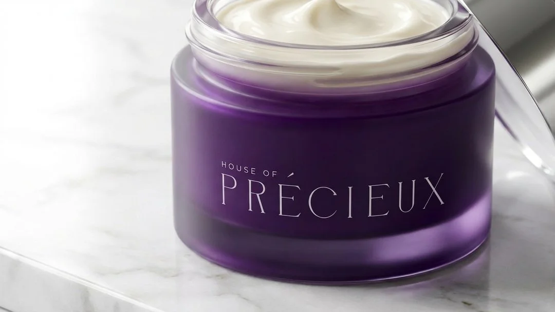 A jar of House of Précieux cream with a purple container, open lid, and visible cream inside, placed on a white marble surface.