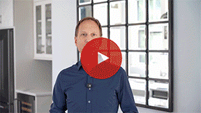 A man in a blue shirt standing indoors near a window with a city view.