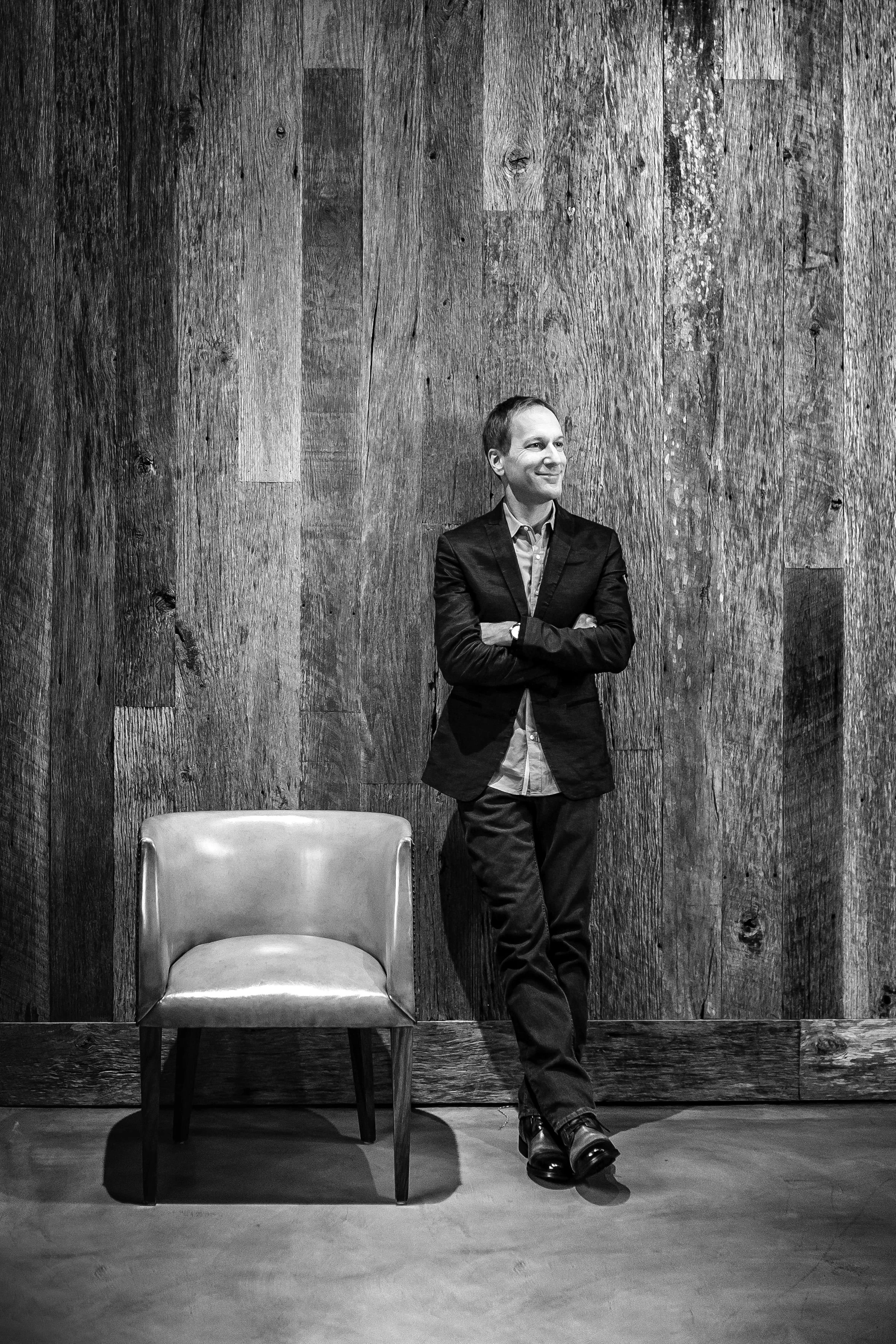 A man in a suit with crossed arms, smiling, standing next to a wooden wall. There is a single chair in front of him.