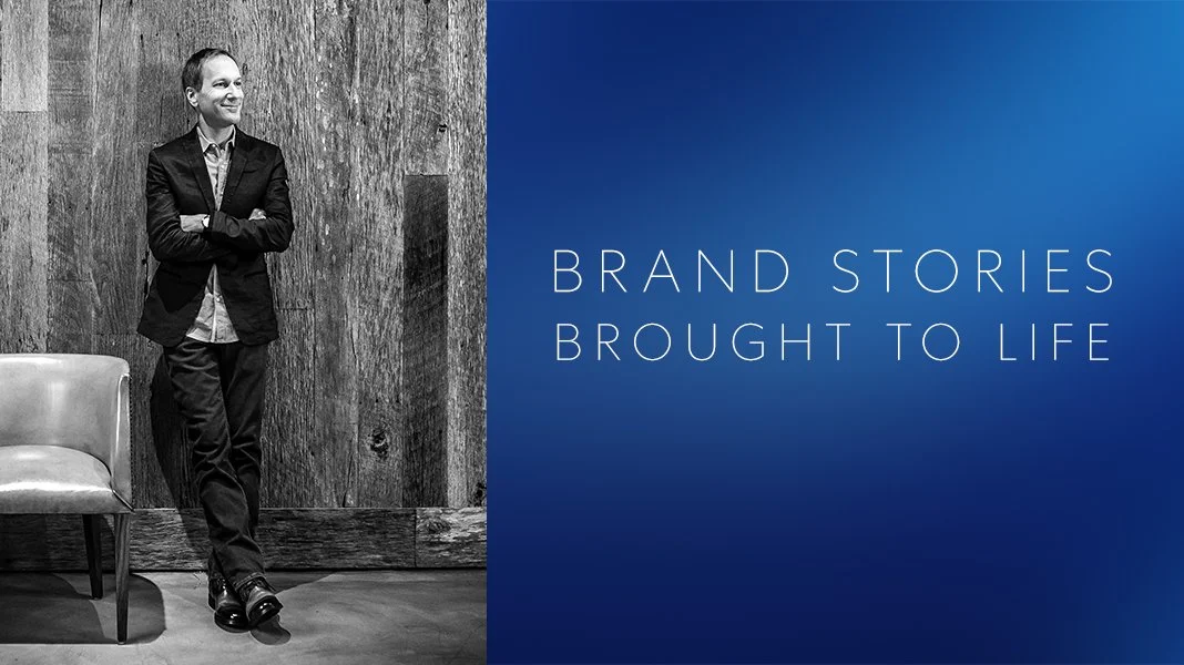 A woman in a suit standing with arms crossed, smiling, in front of a wooden wall, next to a chair, with a blue background and the text 'Brand Stories Brought to Life'
