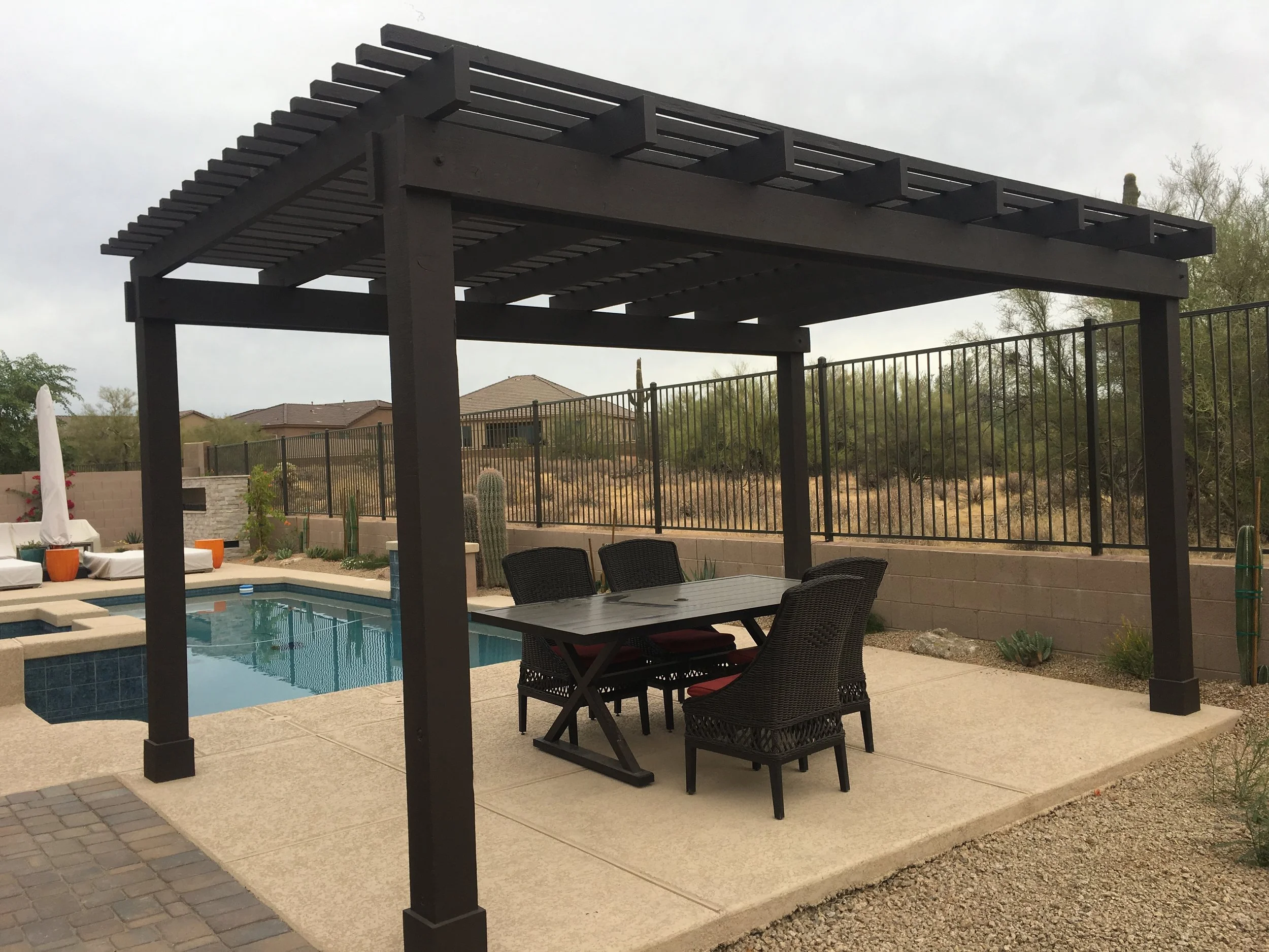 Black pergola over outdoor dining table and chairs near a swimming pool in a backyard with desert landscaping by Blooming Valley Landscape