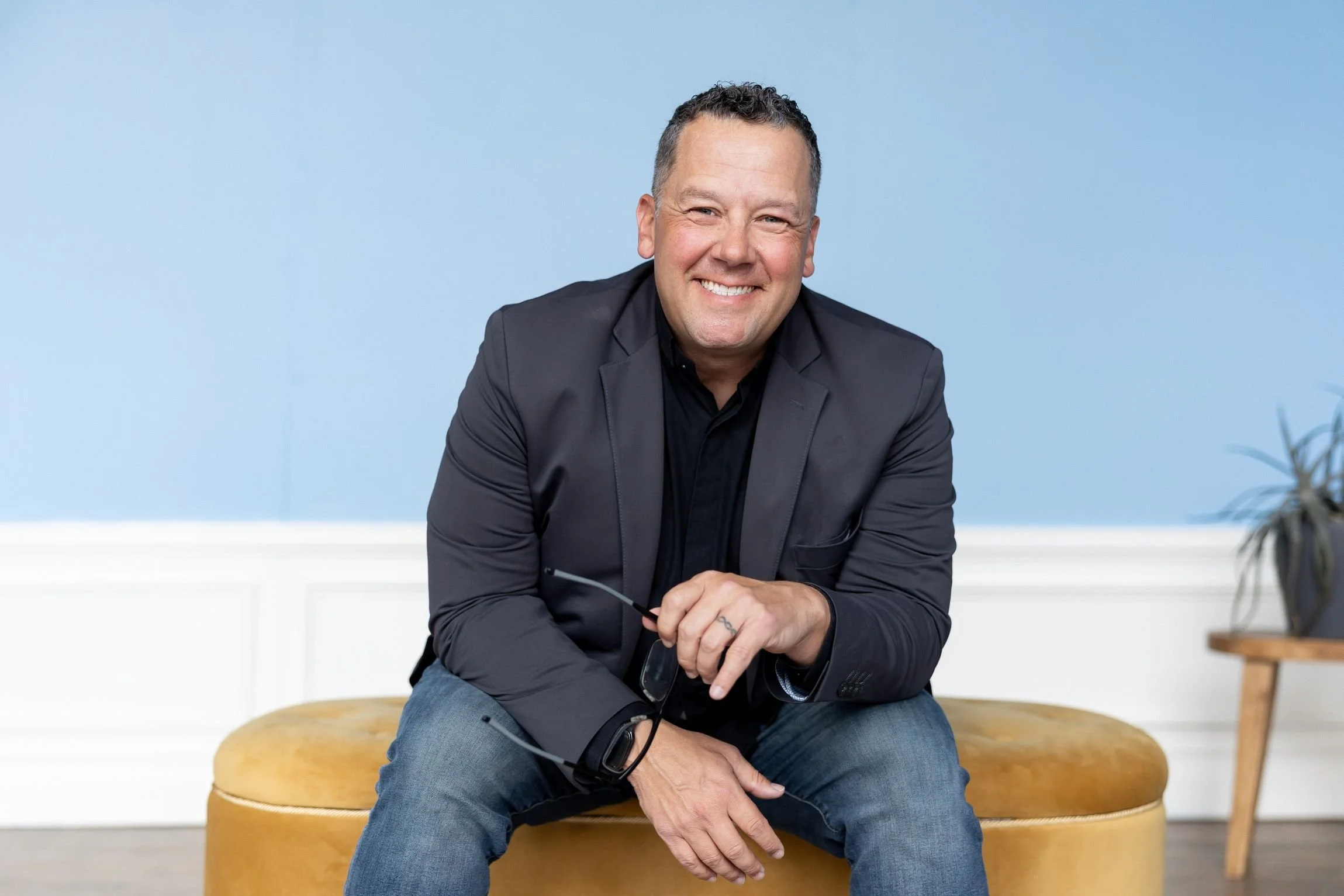 Tony Miltenberger in a black blazer and jeans sitting on a mustard-colored ottoman in front of a light blue wall, holding reading glasses in one hand near his knee, with a potted plant on a wooden stand to the right.