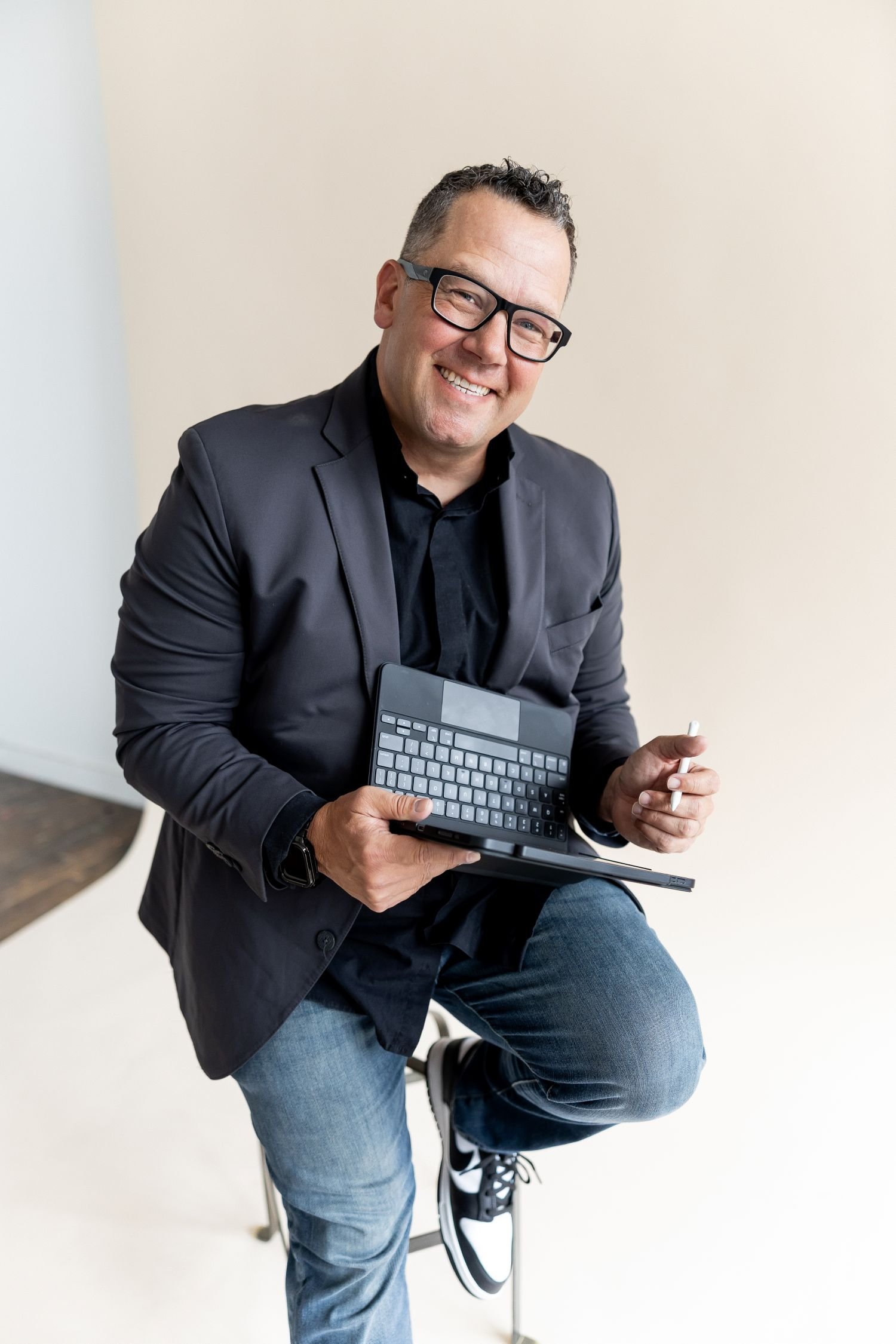Tony Miltenberger with glasses in a black blazer and jeans, holding a small electronic device with a keyboard and a stylus, sitting on a stool against a plain beige wall.