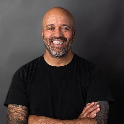 Smiling man with a shaved head and beard, wearing a black t-shirt, standing arms crossed against a gray background.