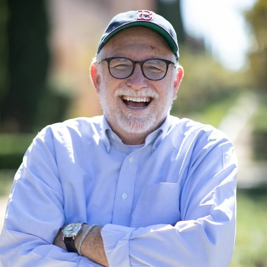 Smiling elderly man with glasses, white beard, wearing a Boston Red Sox cap and a light blue button-up shirt, outdoors on a sunny day.