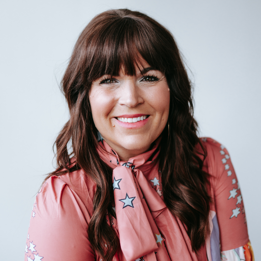 A woman with dark brown hair and bangs smiling, wearing a pink blouse with star patterns and a matching star-patterned scarf.