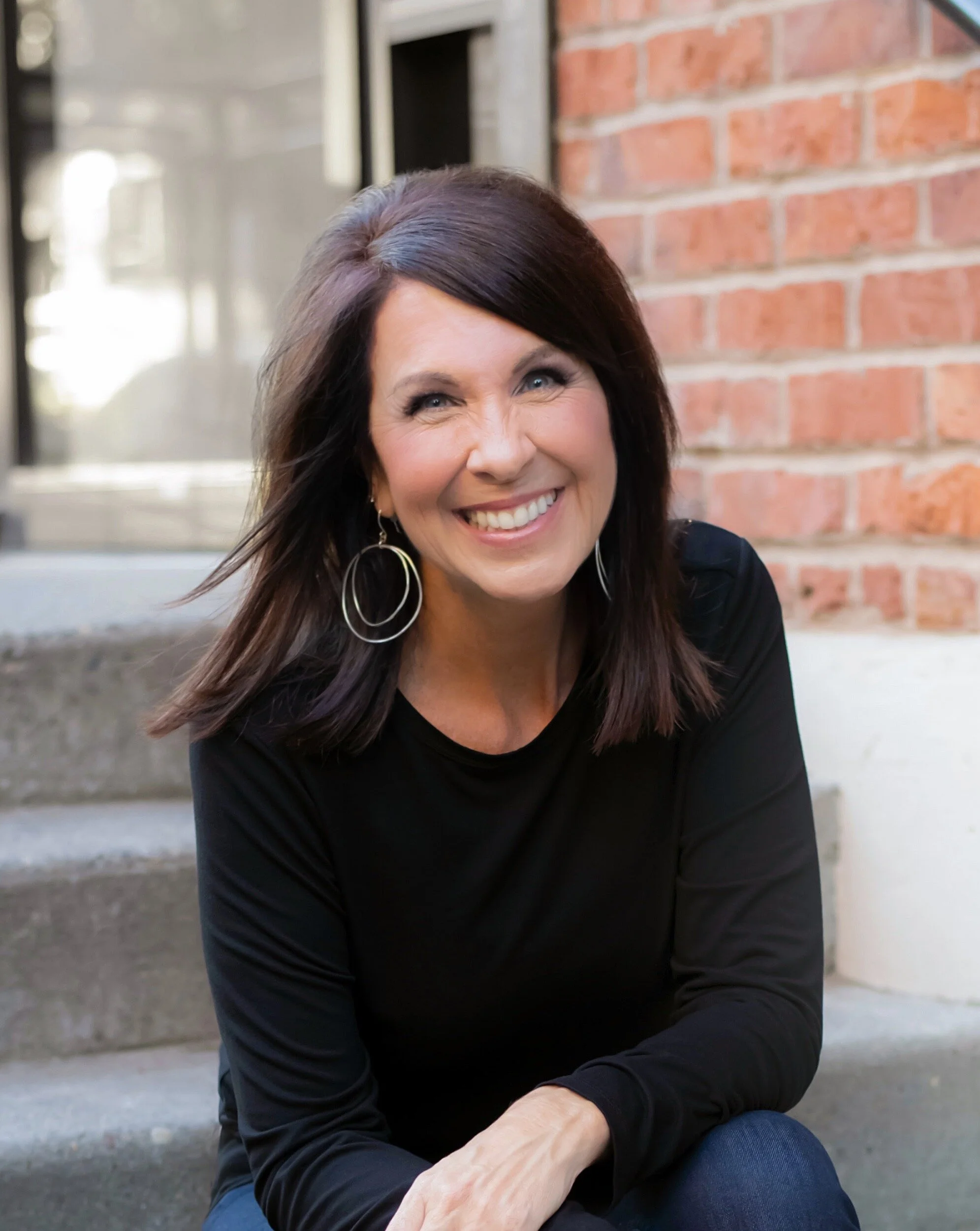 A woman with shoulder-length dark hair, wearing a black top and large hoop earrings, smiling while sitting on outdoor steps in front of a brick building.