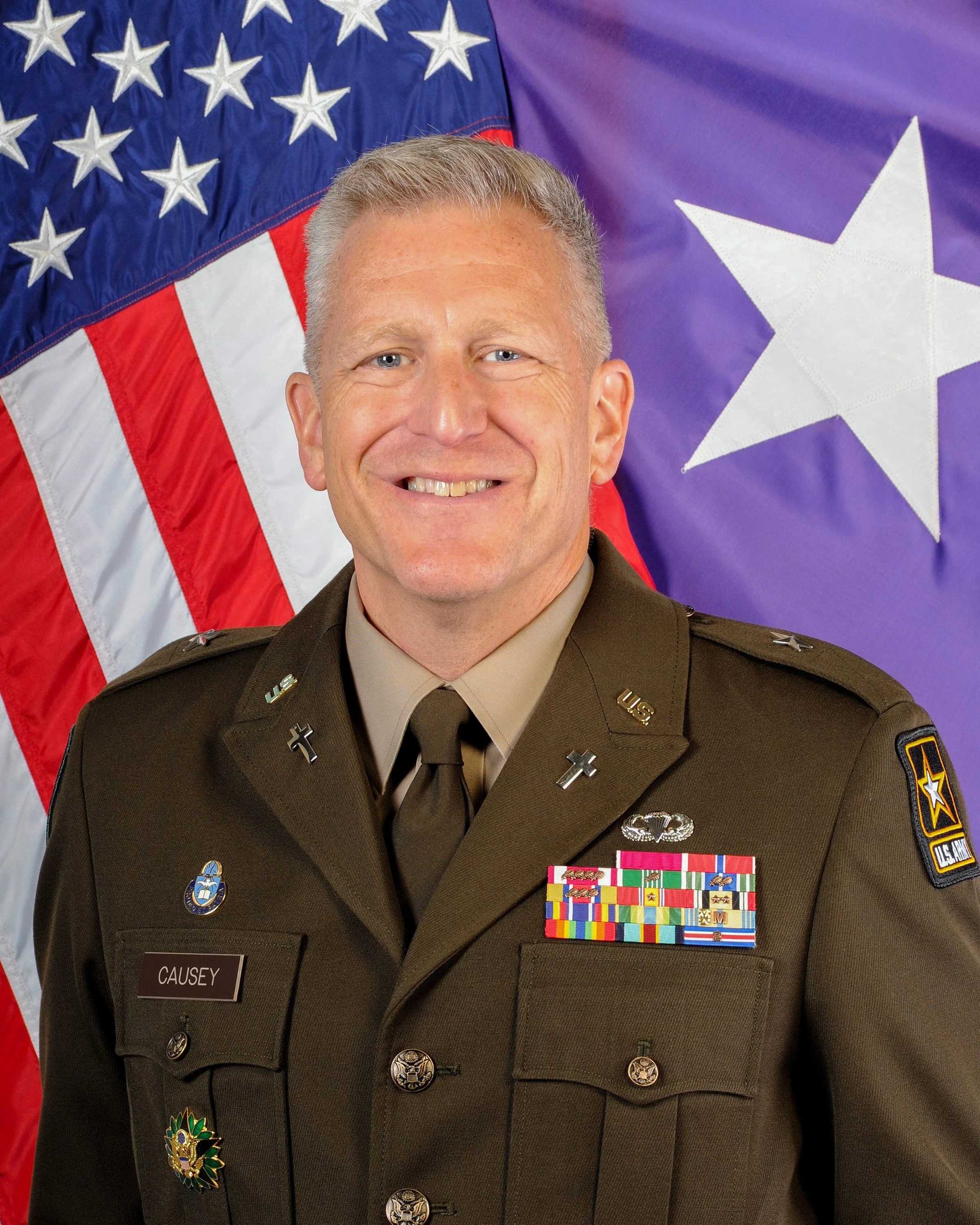 A smiling man in a U.S. Army uniform standing in front of an American flag and a purple flag with a white star.
