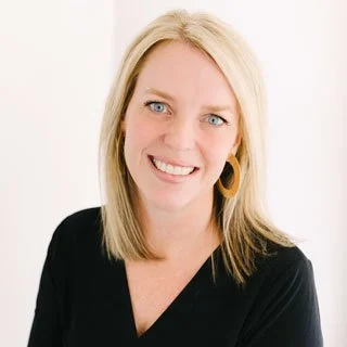 Portrait of a smiling woman with blonde hair, wearing a black top and gold hoop earrings, against a light background.