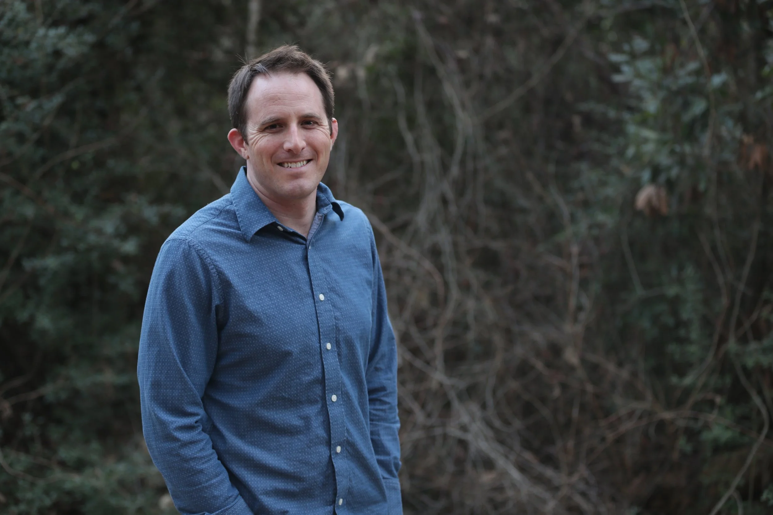 A man in a blue button-up shirt standing outdoors in front of a background of bushes and trees.