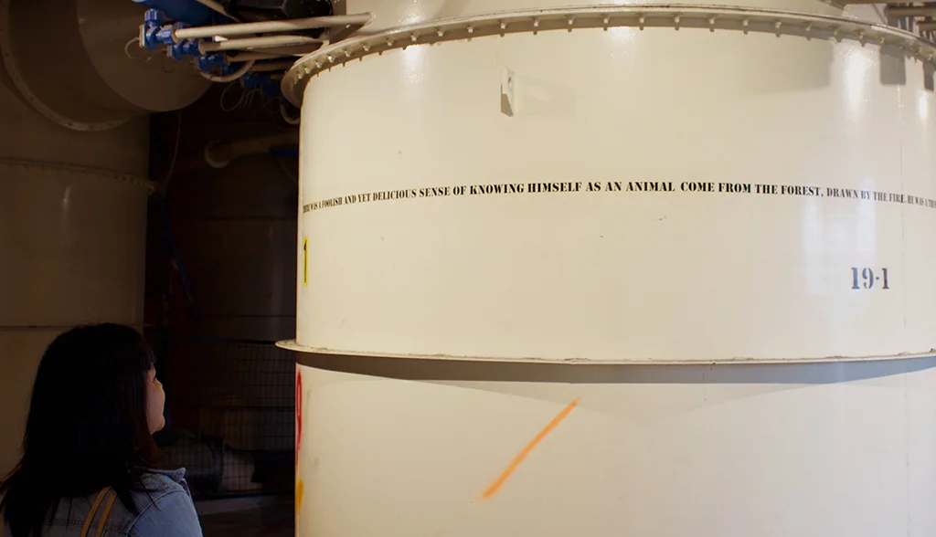 He was a thing , Spray paint text installation on factory drum, sound installation (cattle lowing), 2017. He was a thing beckons viewers to circumambulate an industrial drum, filled with remnant detergent material from the bygone days of the Unilever factory. On the cylindrical form is an excerpt from Ray Bradbury's Fahrenheit 451 (1953): THERE WAS A FOOLISH AND YET DELICIOUS SENSE OF KNOWING HIMSELF AS AN ANIMAL, COME FROM THE FOREST, DRAWN BY THE FIRE. HE WAS A THING OF BRUSH AND LIQUID EYE, OF FUR, AND MUZZLE AND HOOF, HE WAS A THING OF HORN AND BLOOD, THAT WOULD SMELL LIKE AUTUMN IF YOU BLED IT OUT ON THE GROUND.
