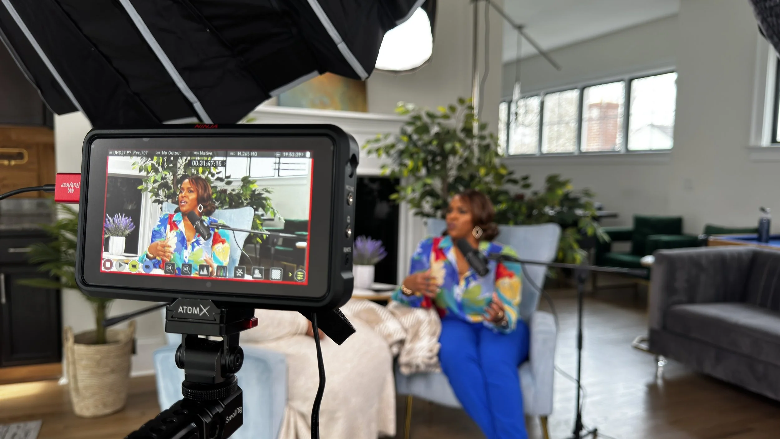 A woman in a colorful blouse and blue pants is speaking into a microphone during an interview or recording, with a camera capturing her image on its screen, inside a bright room with large windows and green plants.