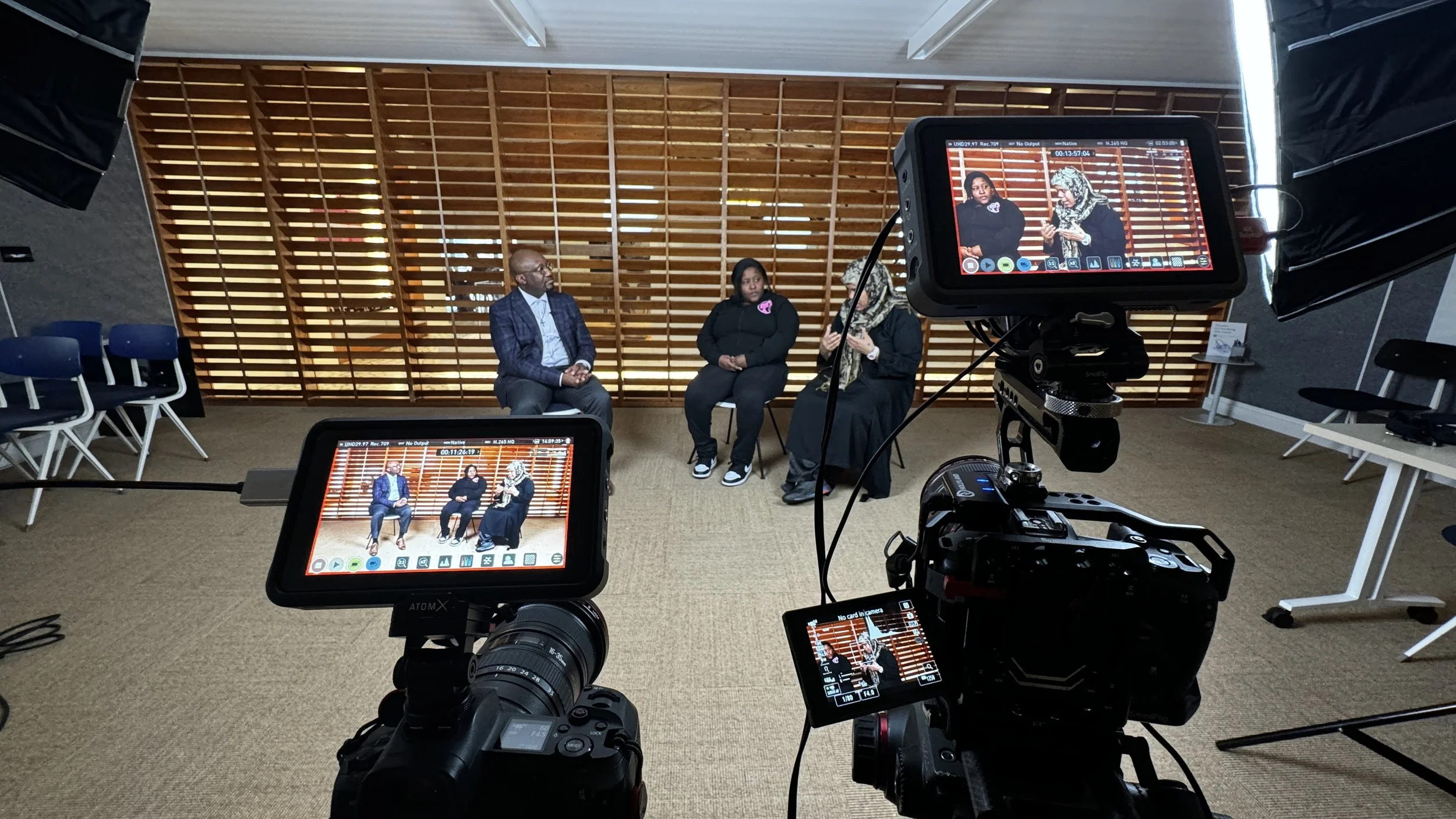 Three people seated on stage during a recorded panel discussion or interview, with two cameras capturing the scene in a room with a wooden slat wall background and a beige carpet.